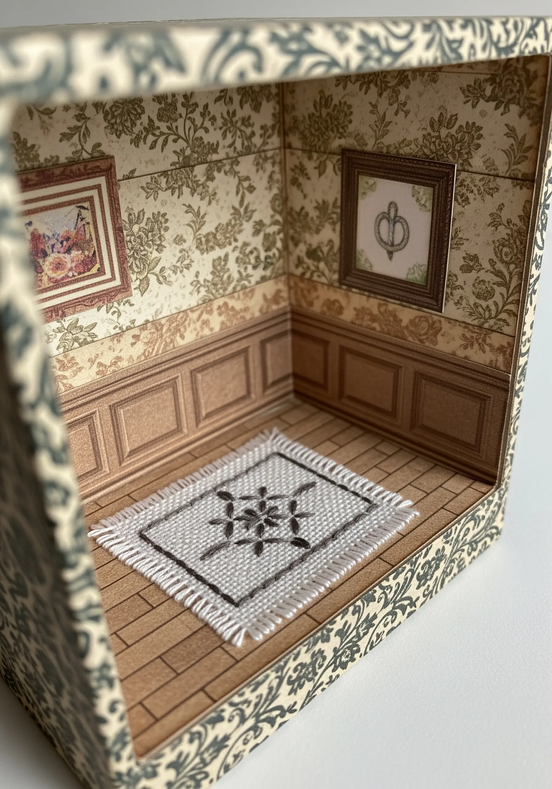A miniature room diorama in a box, featuring a tiny woven rug made from embroidery floss.