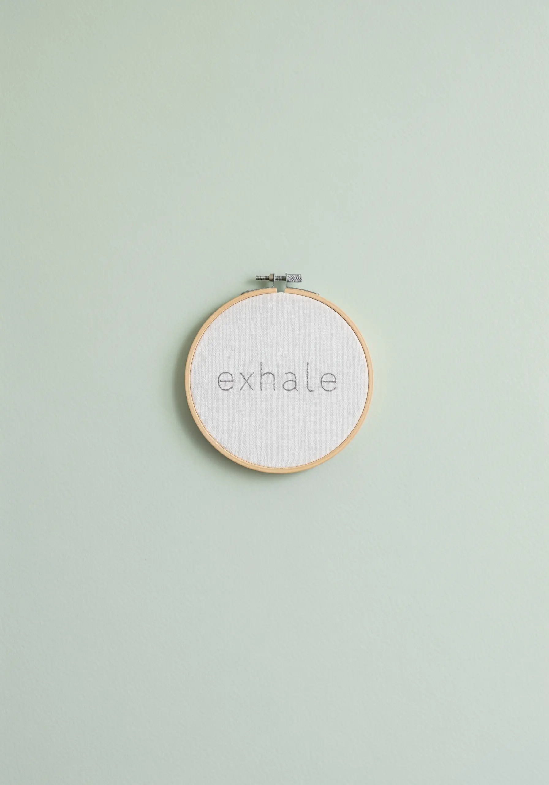 The word 'exhale' in tiny, delicate grey cross-stitch on white Aida cloth.