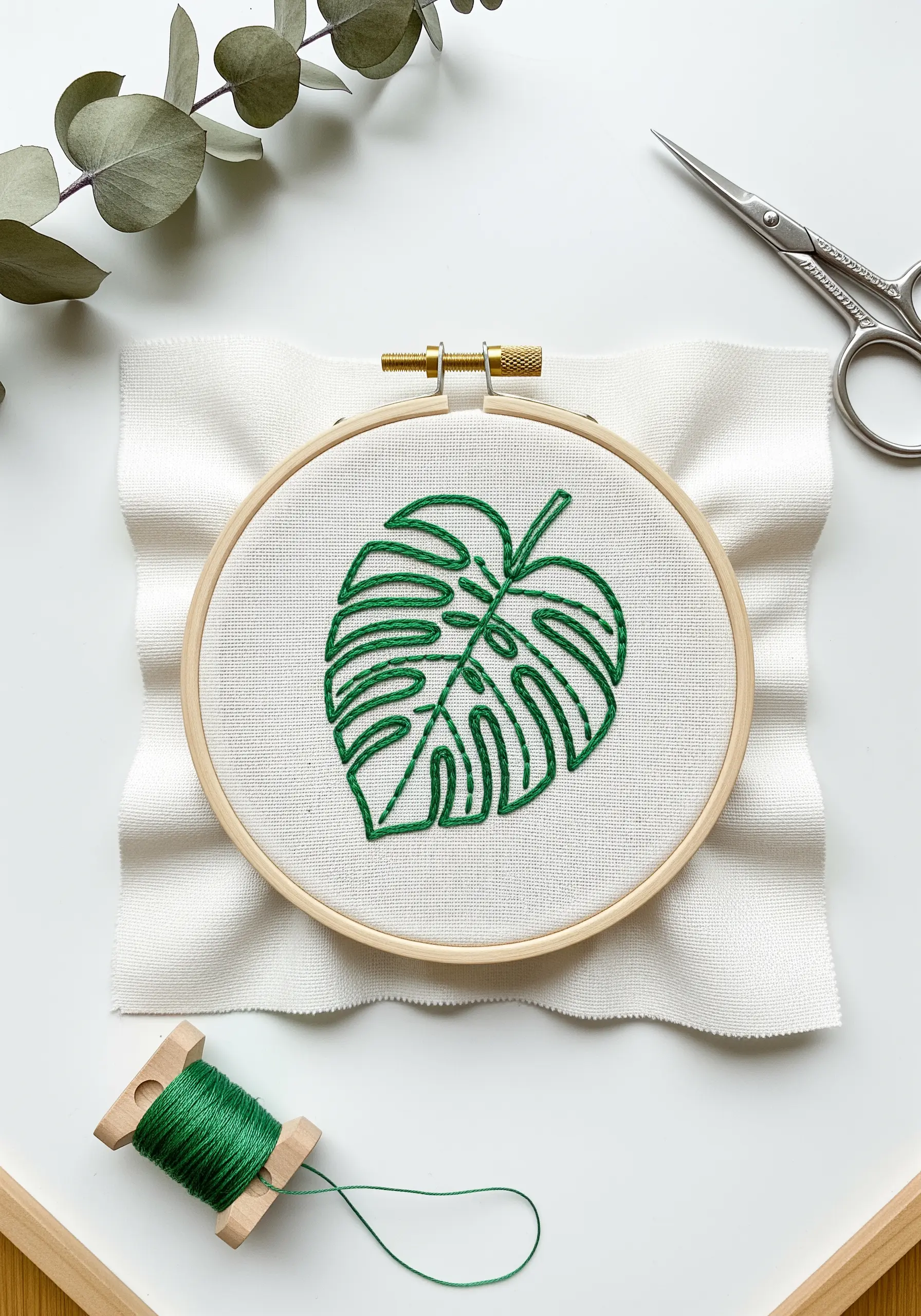 A minimal outline of a monstera leaf embroidered in green thread on off-white fabric.