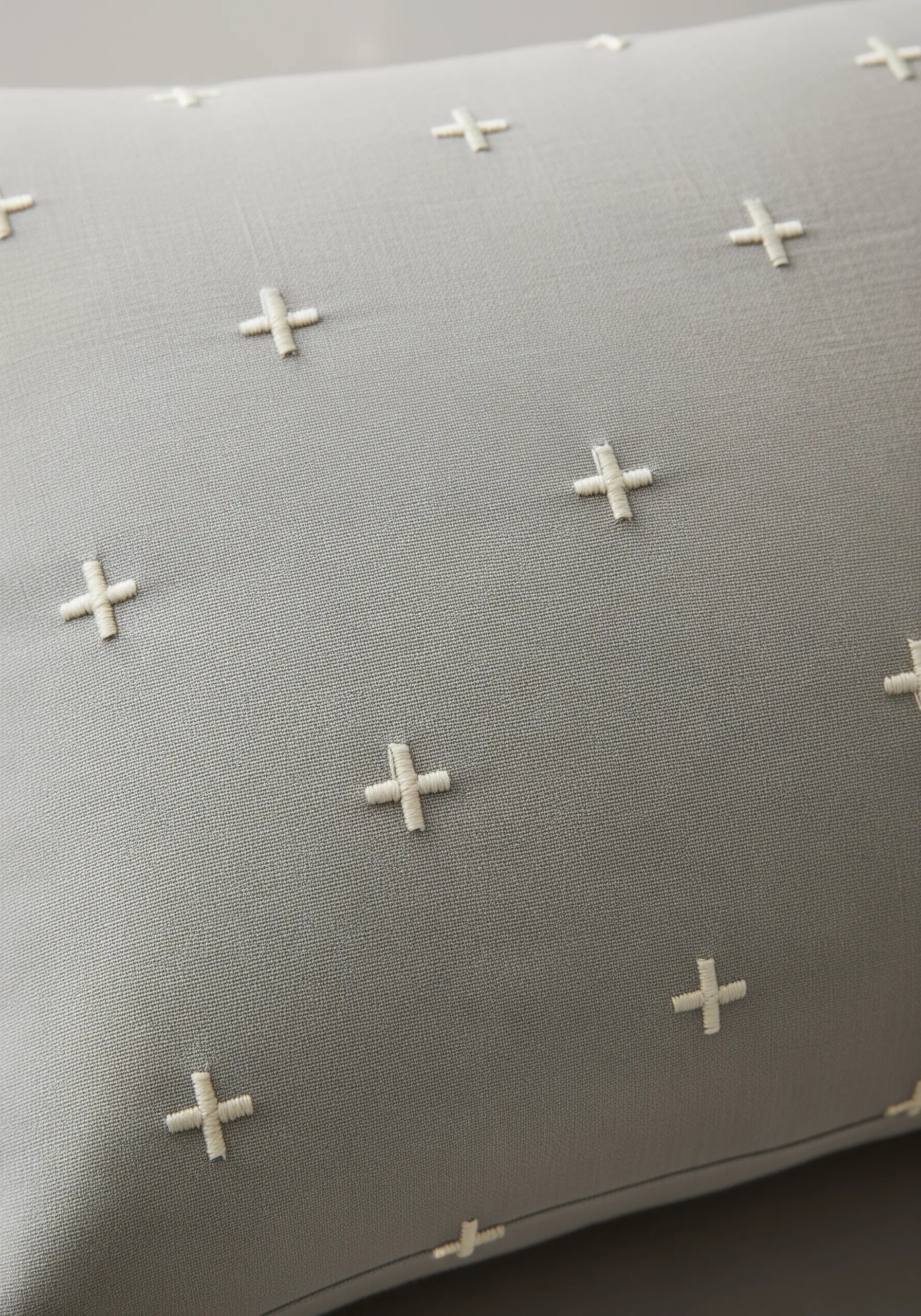 A minimalist geometric pattern of small, embroidered off-white plus signs on a grey pillow.