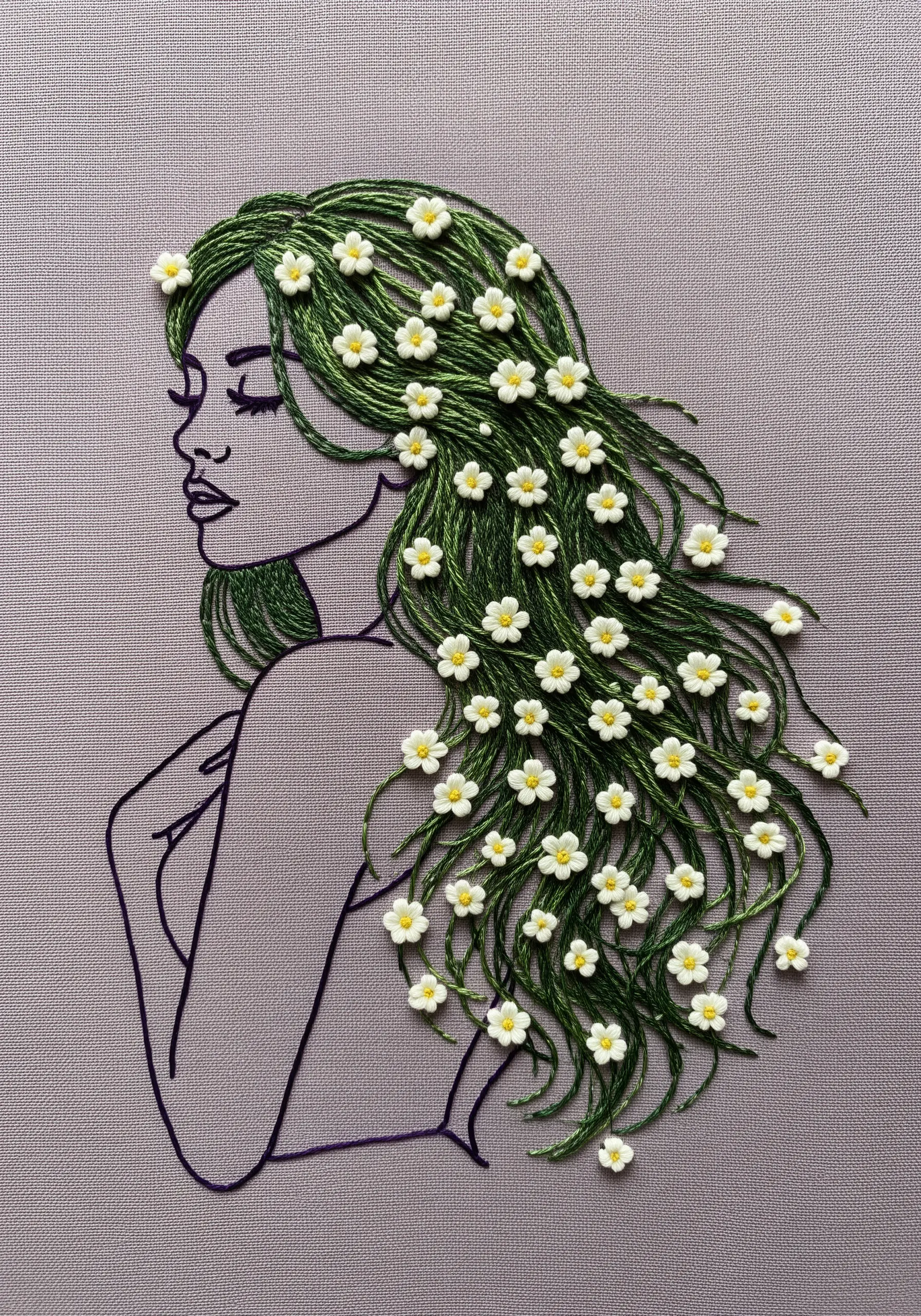 A line-art embroidery of a woman's profile, with her long green hair filled with tiny daisies.