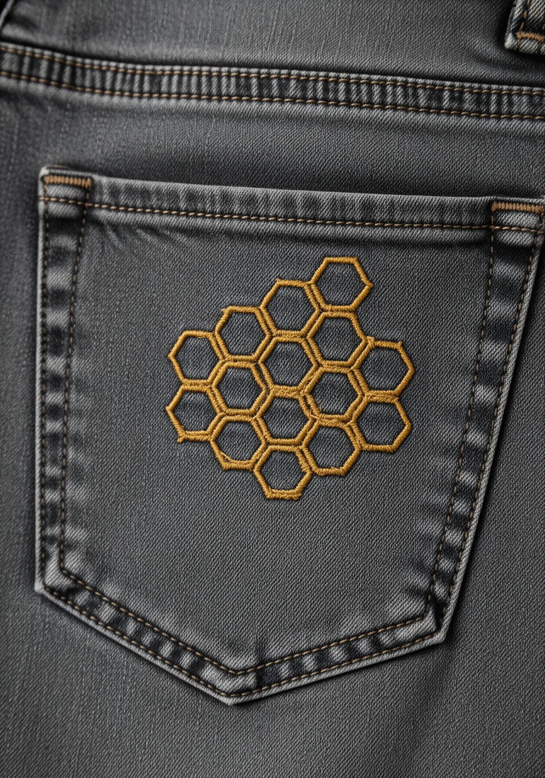 A geometric honeycomb pattern stitched in gold thread on a charcoal grey jean pocket.