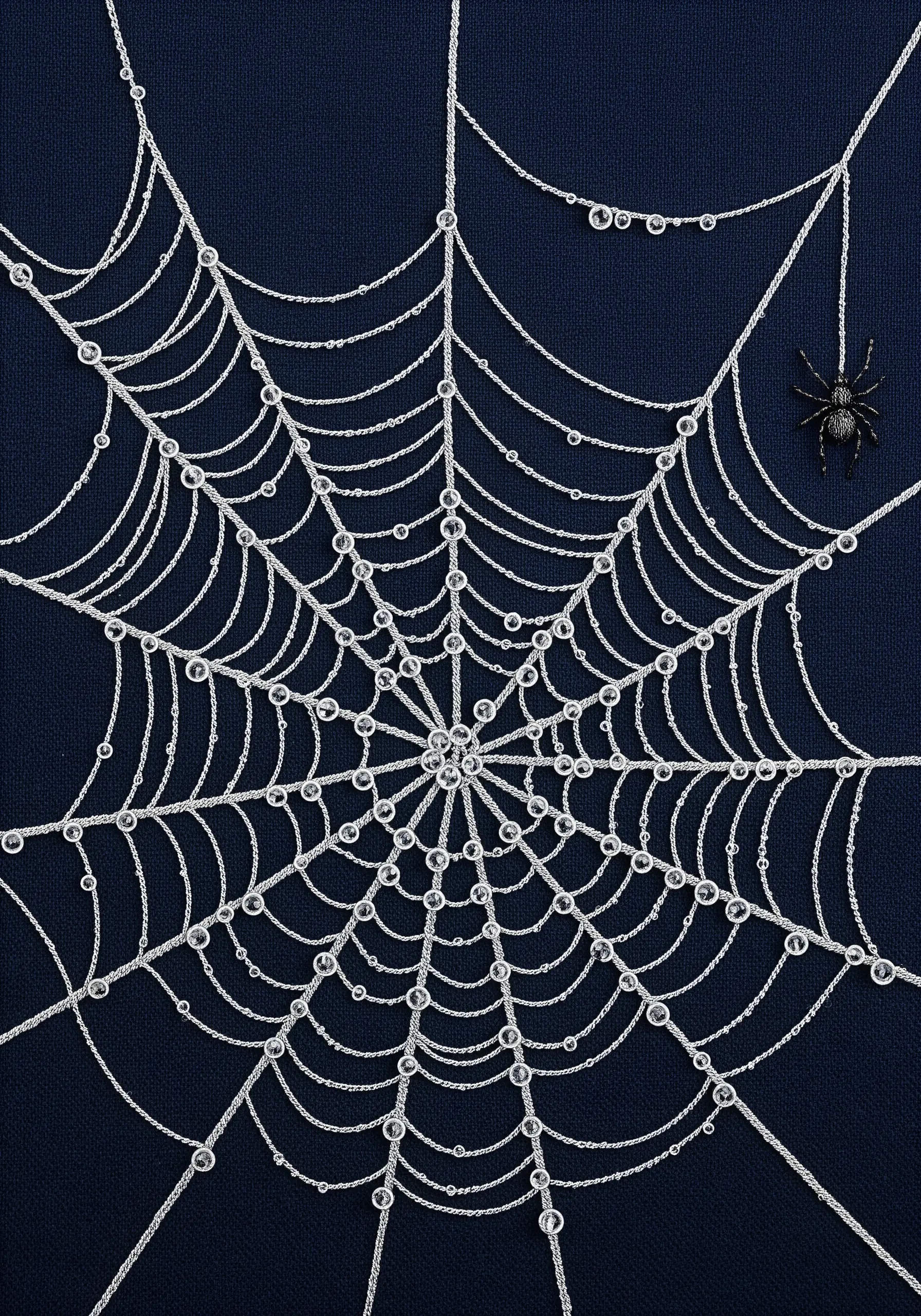 An intricate silver spiderweb on dark blue fabric, adorned with tiny beads that look like dewdrops.