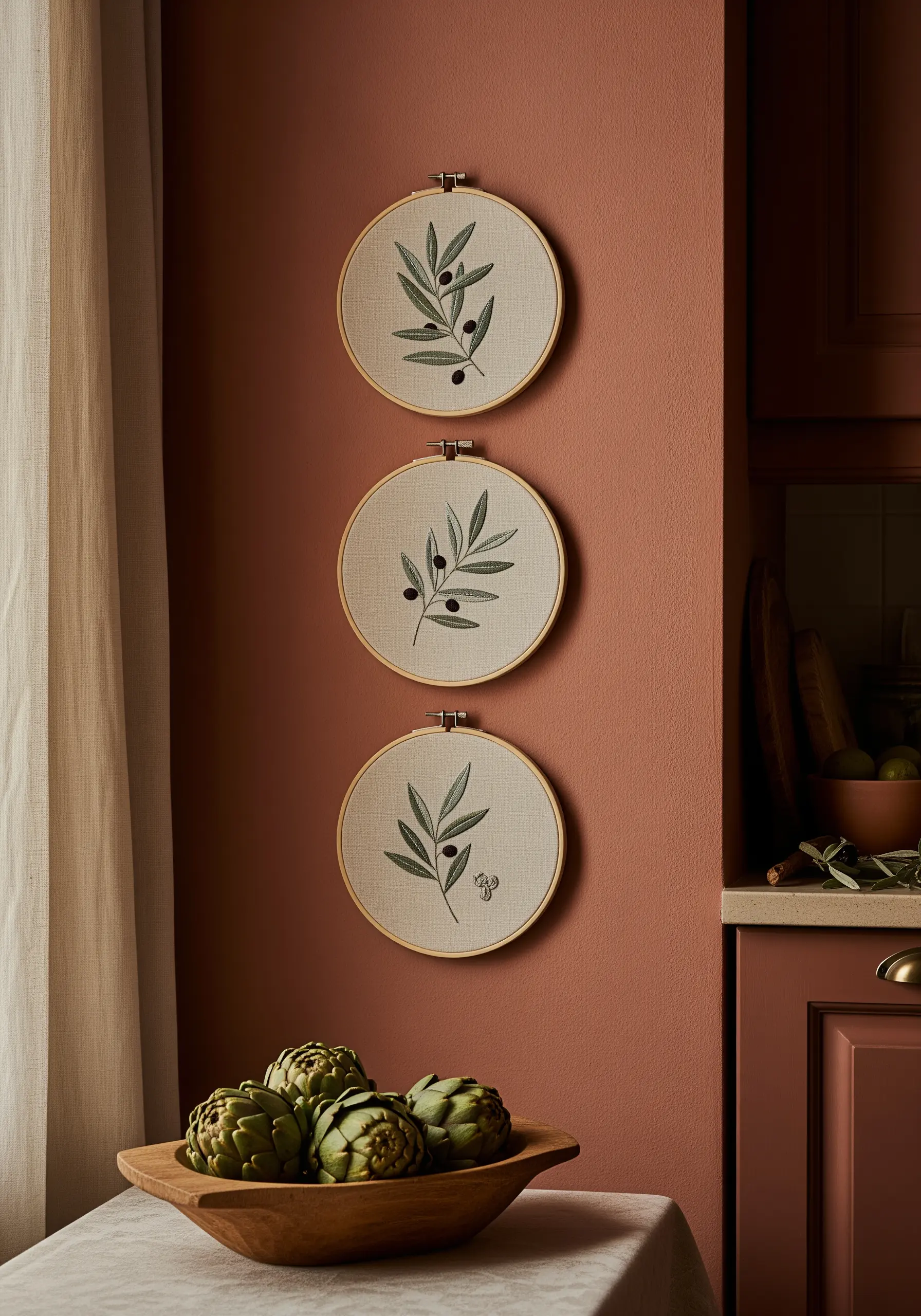 Three vertically hung embroidery hoops, each with a minimal olive branch design against a terracotta wall.