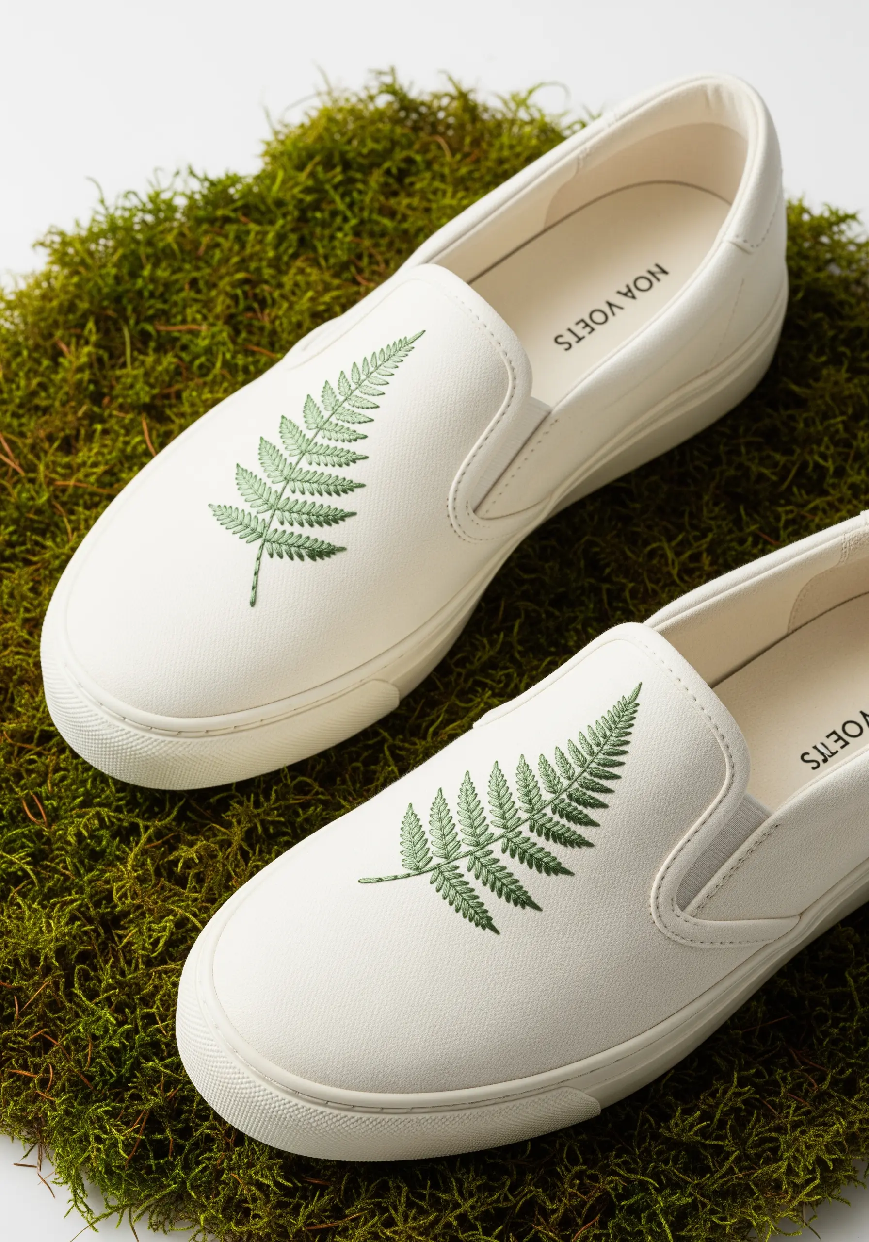 A single, delicate fern leaf embroidered on the top of a white slip-on sneaker.