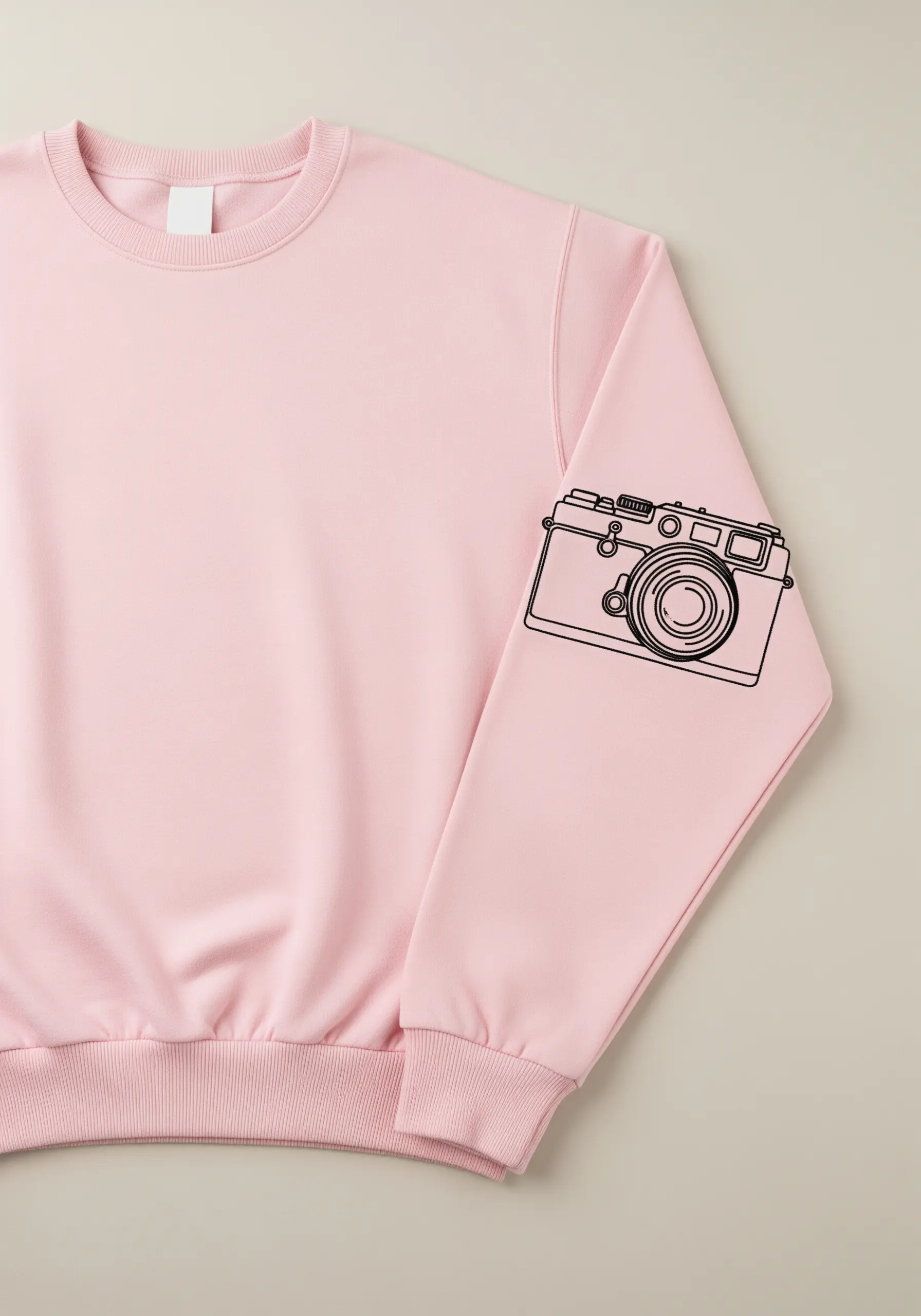 A detailed, fine-line illustration of a vintage camera embroidered in black on a pink sweatshirt sleeve.