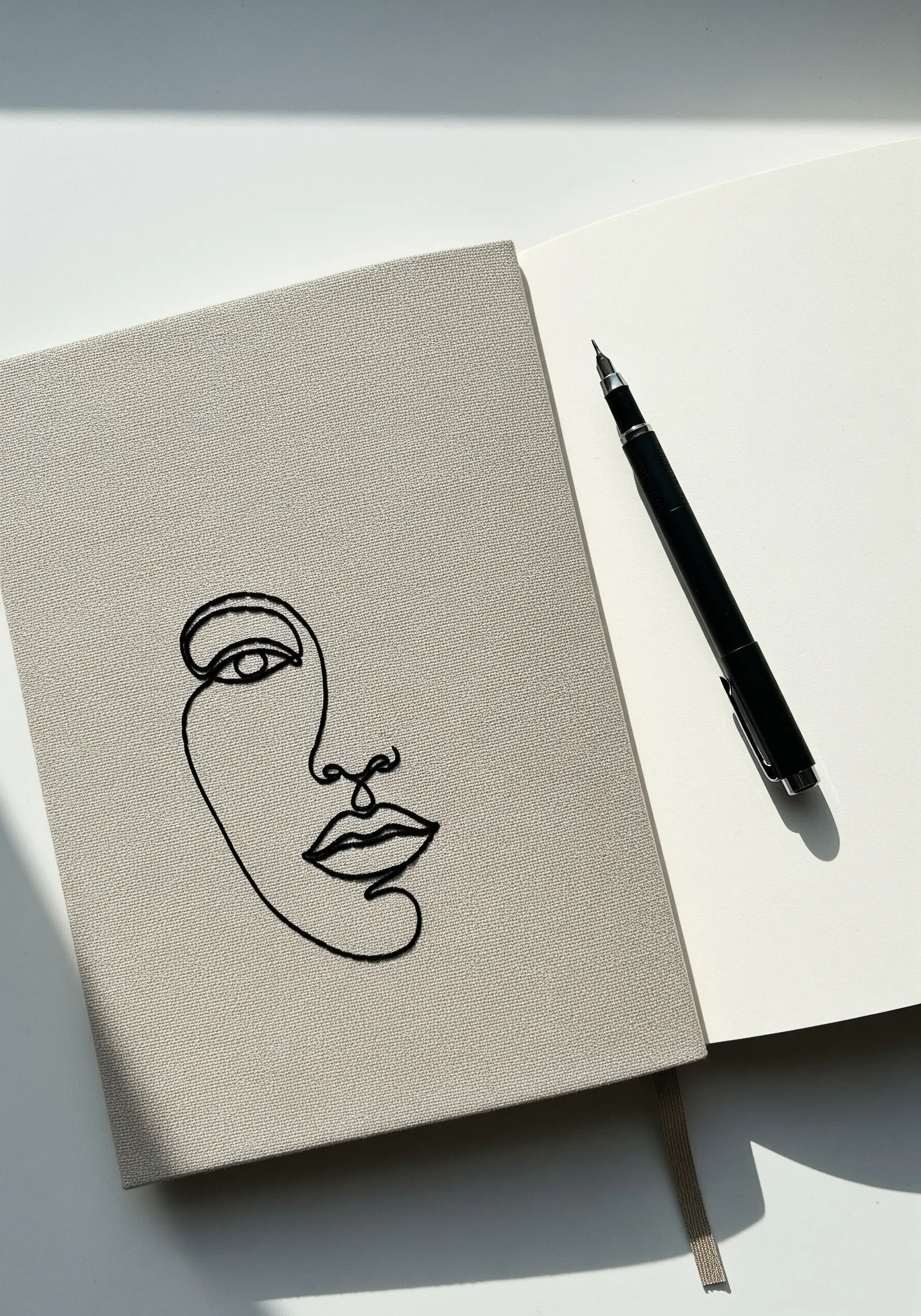 Abstract face drawn in a single, continuous black embroidered line on a grey notebook.