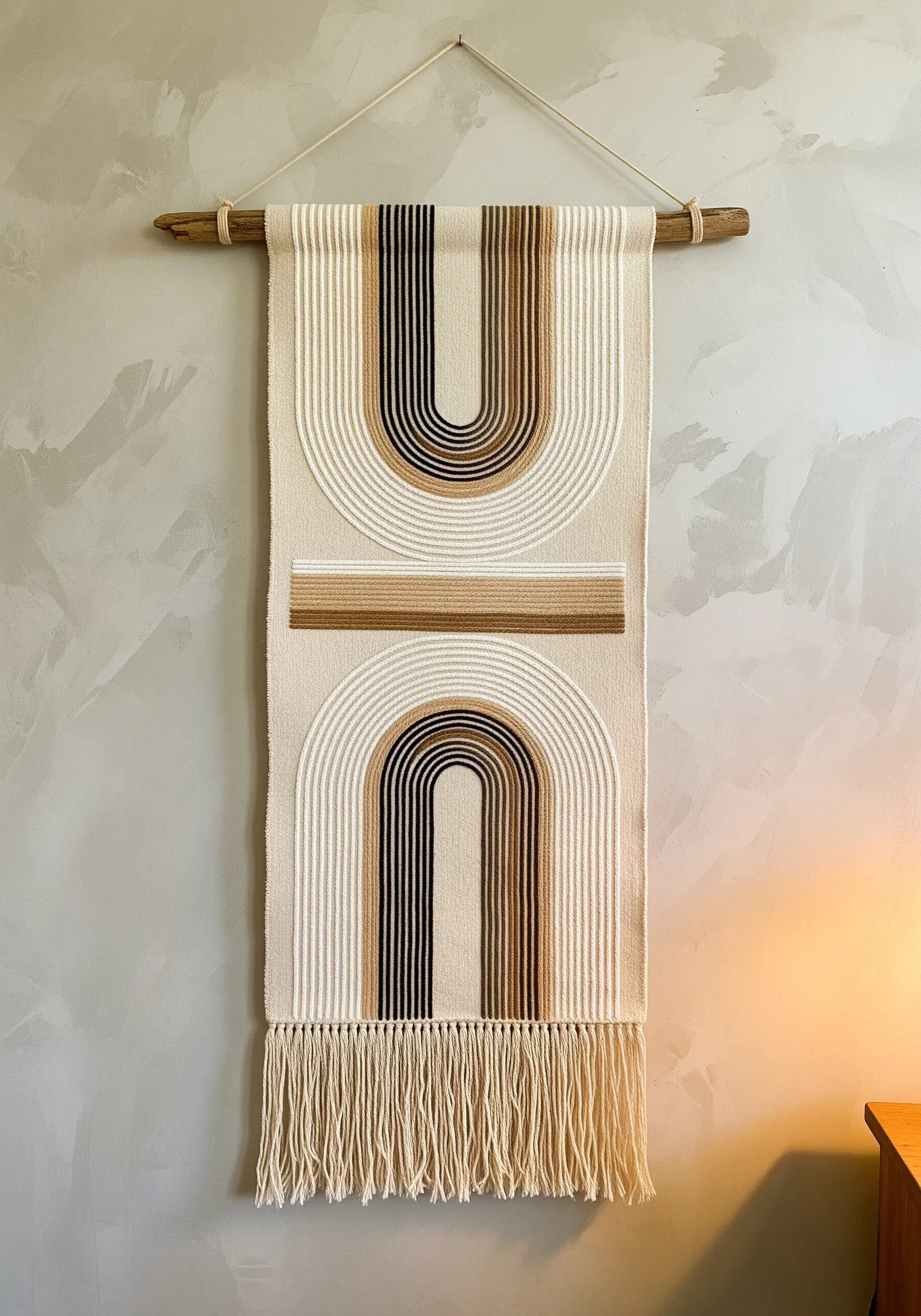 A large woven wall hanging with geometric neutral-toned arches and a fringed bottom.