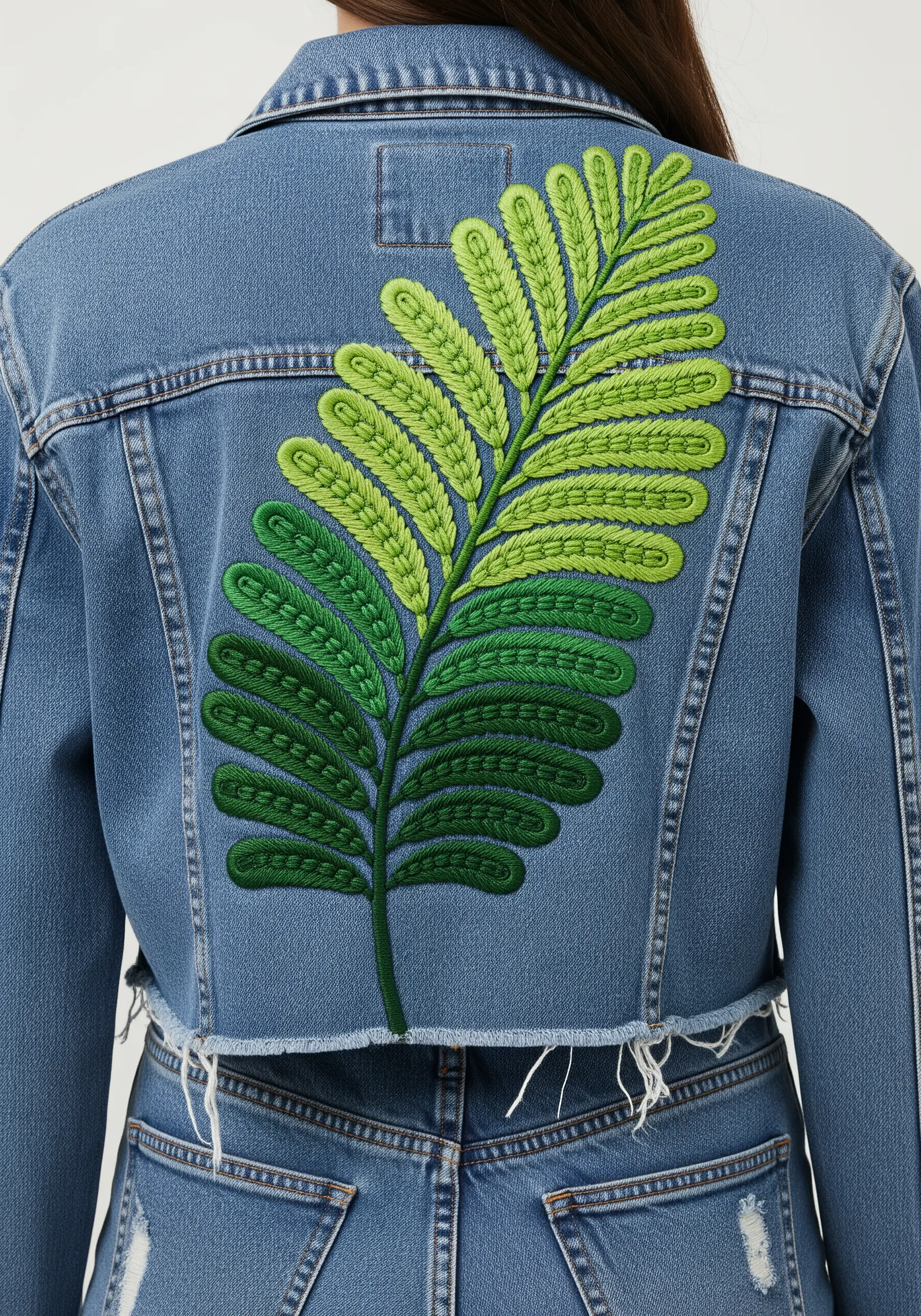 A large fern leaf embroidered on the back of a denim jacket with a dark-to-light green gradient.