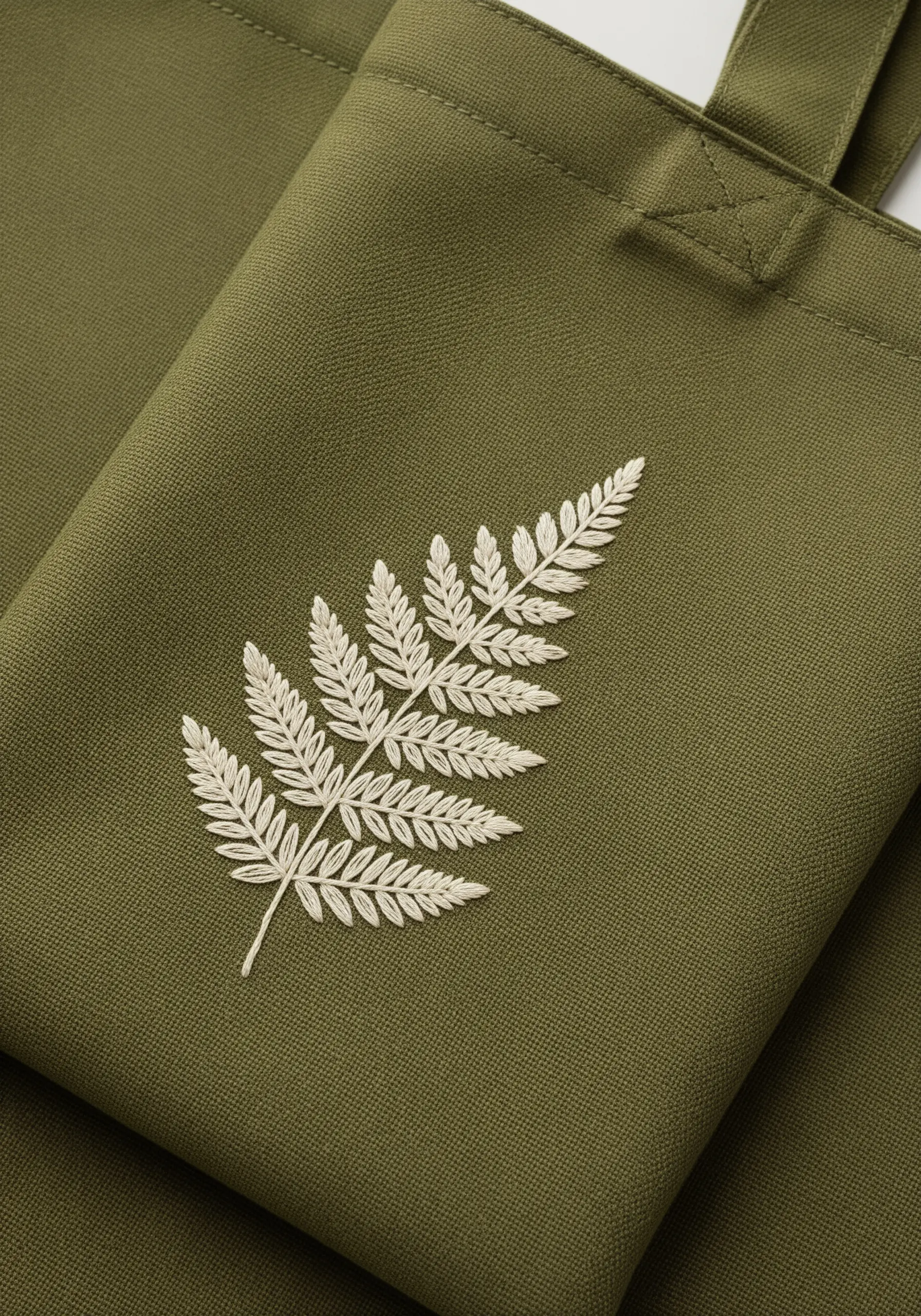 A detailed white fern leaf embroidered on a dark olive green tote bag.