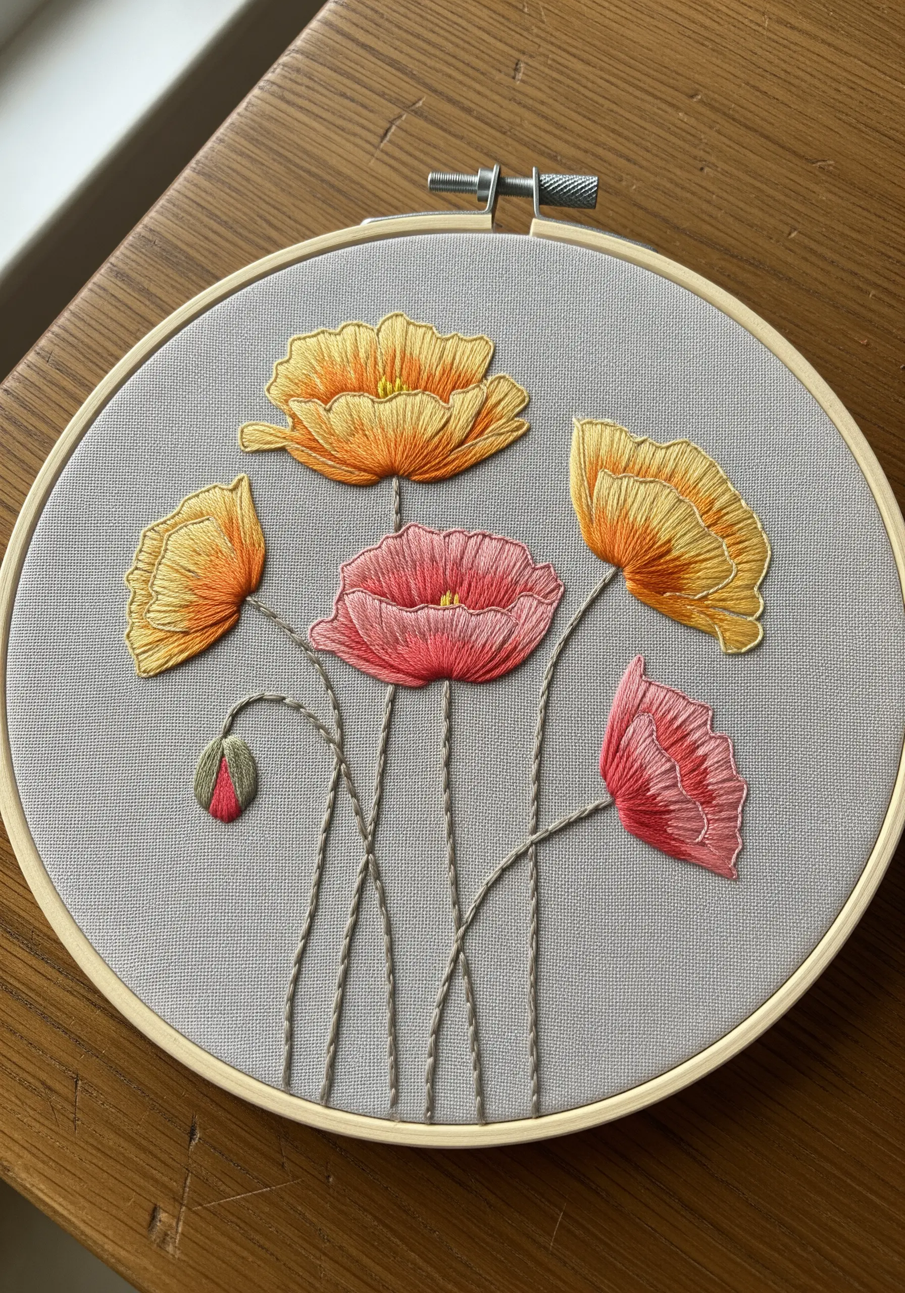 Hand-embroidered poppies with yellow and pink thread blending on grey fabric in a hoop.