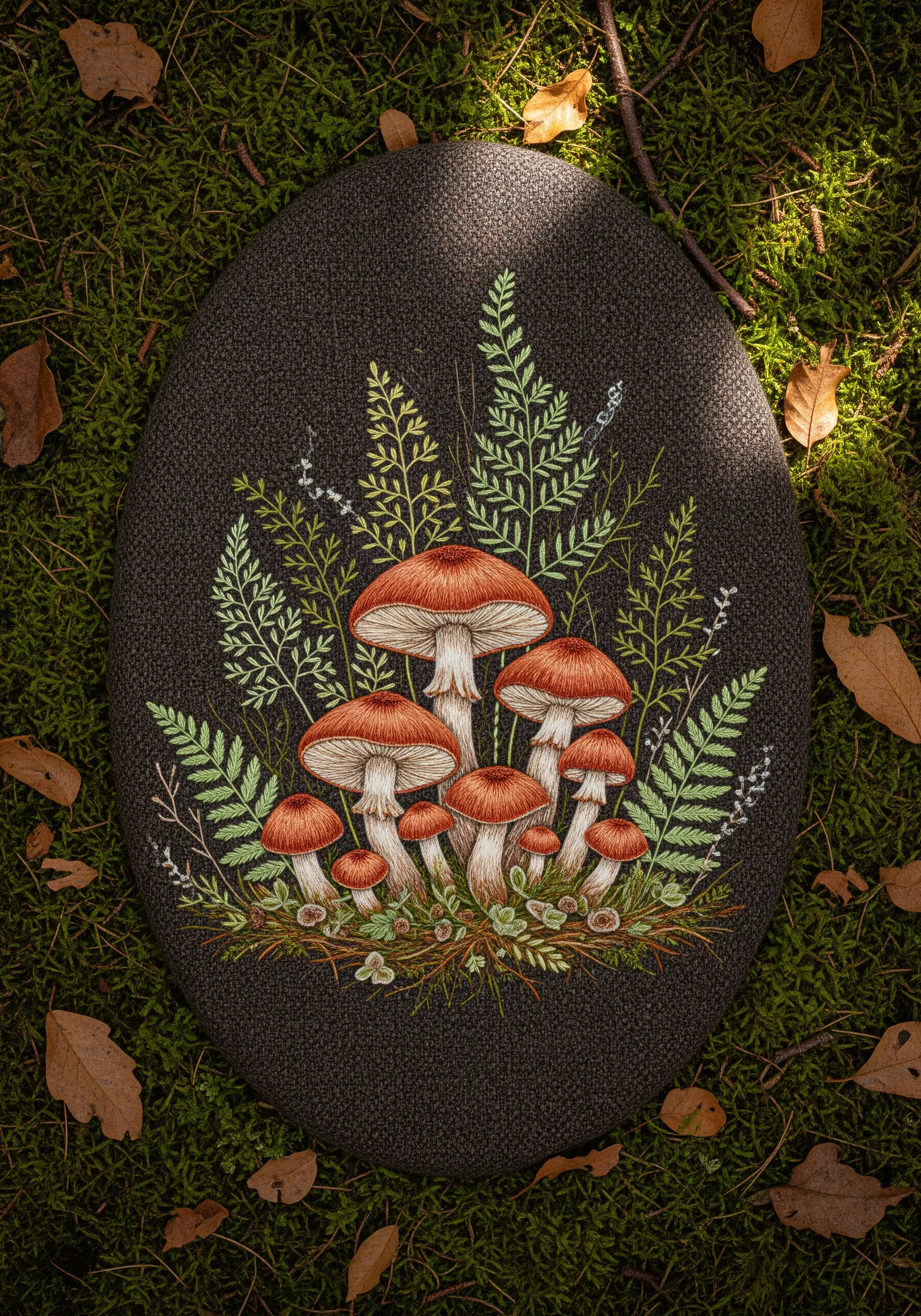 Embroidery of a cluster of detailed brown mushrooms and ferns on dark fabric.