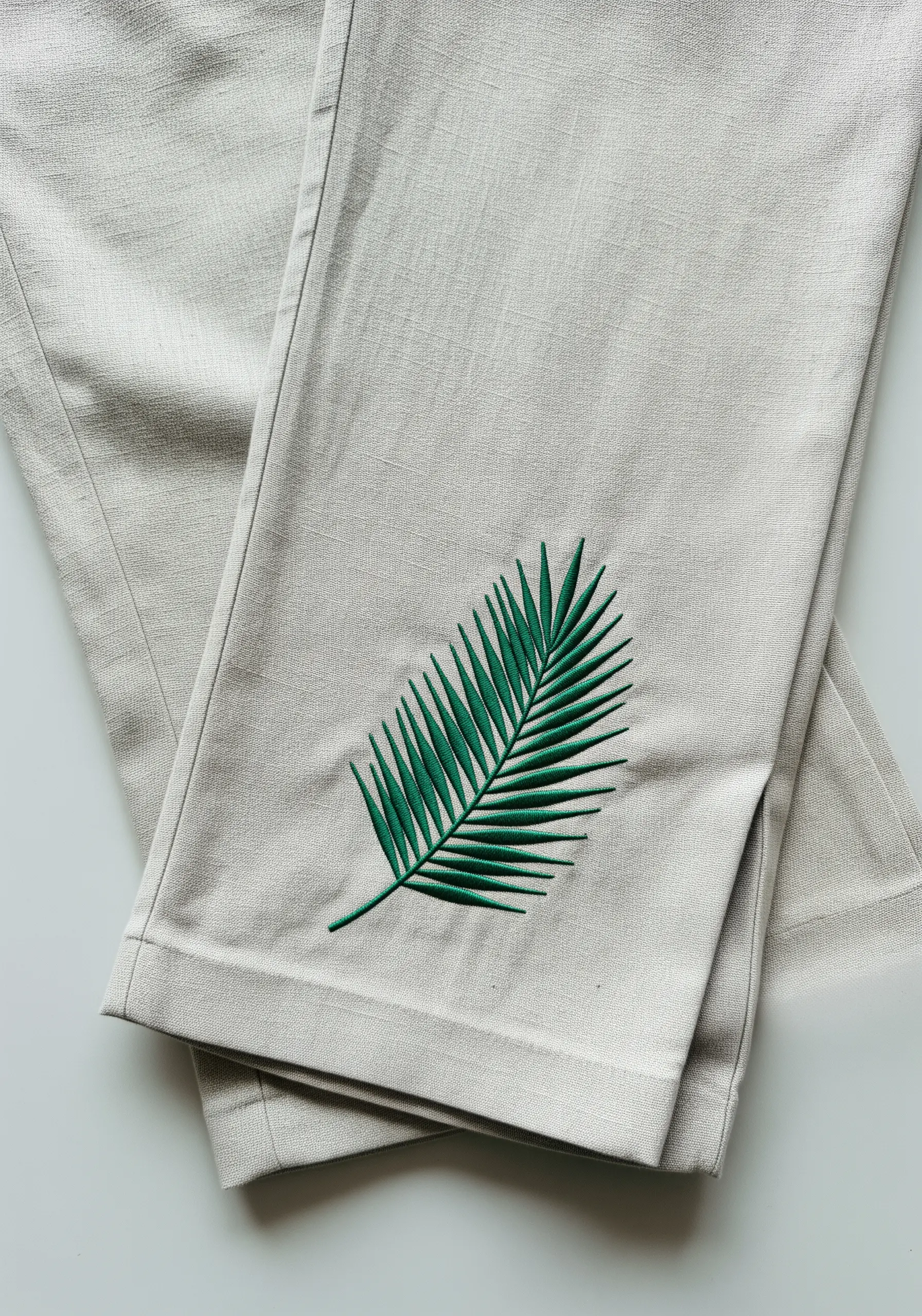 A single green palm leaf embroidered on the hem of light-colored linen pants using fishbone stitch.
