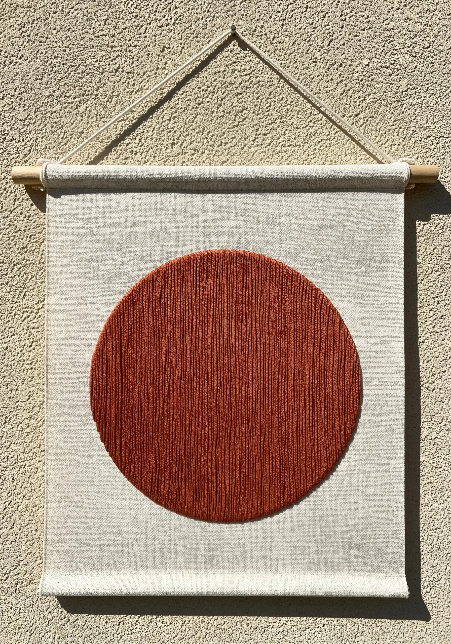 A minimalist terracotta oval filled with vertical couched threads on a white canvas wall hanging.