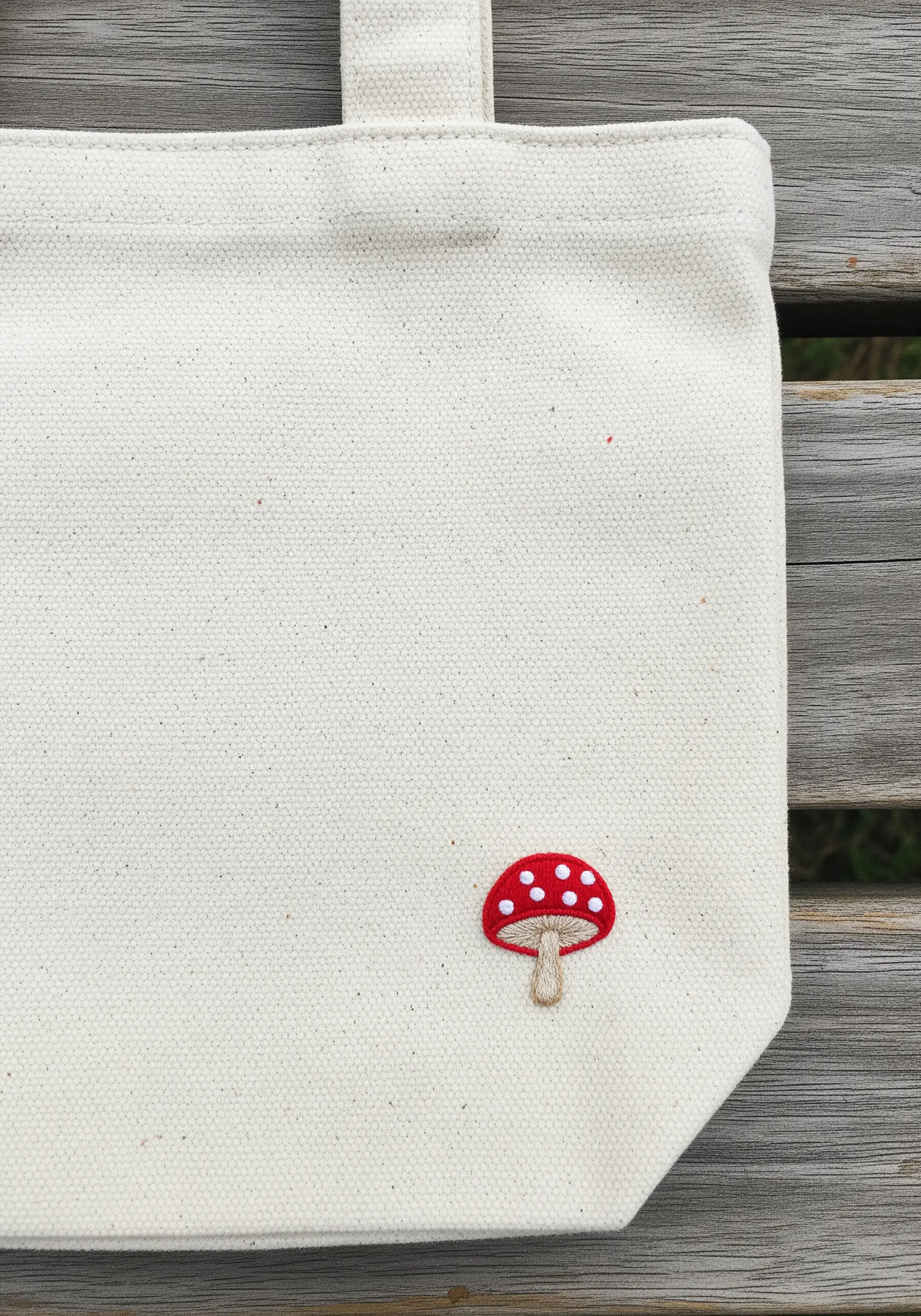 A small red and white mushroom embroidered on the corner of a canvas tote bag.