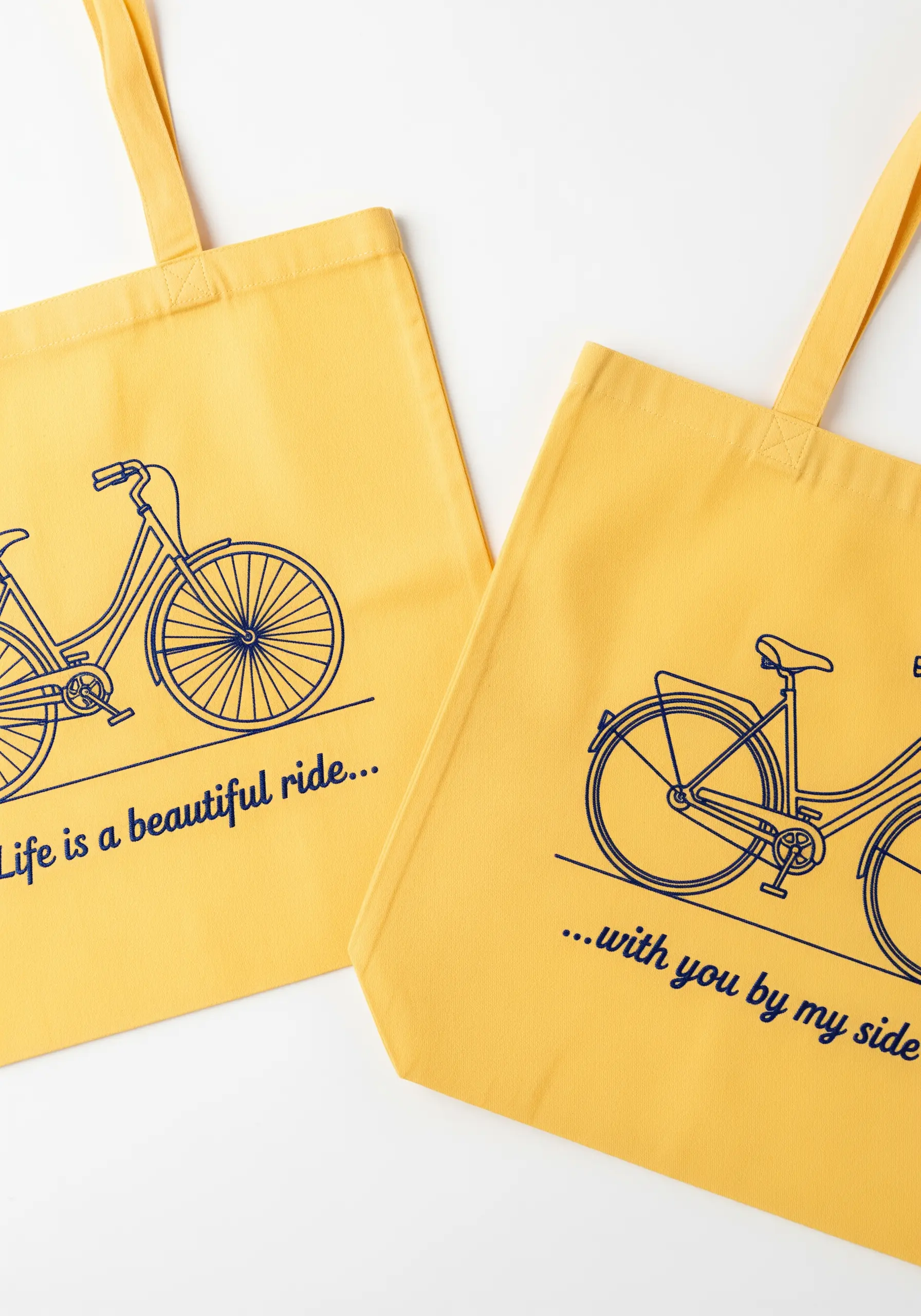 Yellow tote bags with a blue line-art bicycle and a split quote about life's ride.
