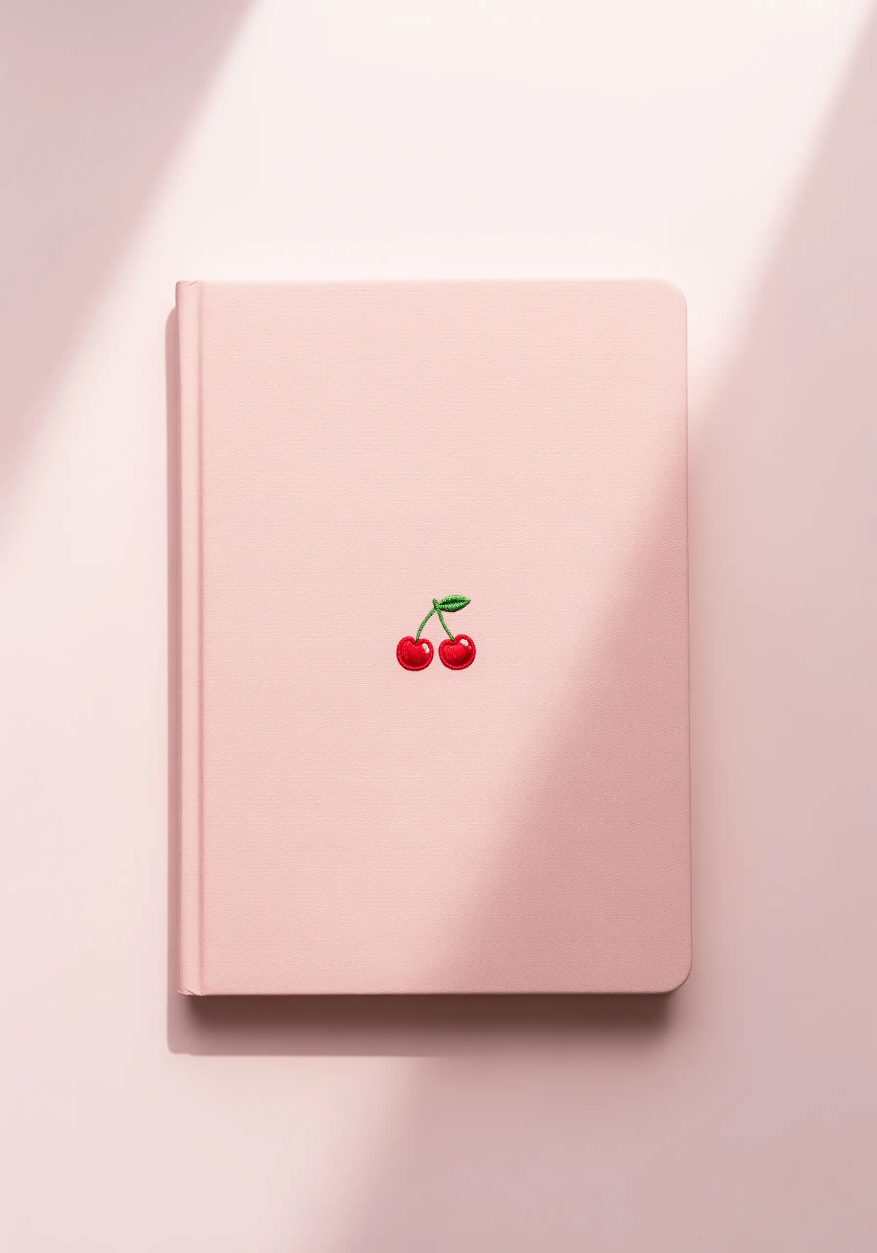 A tiny embroidered cherry motif on a pink notebook cover, with a white highlight stitch.