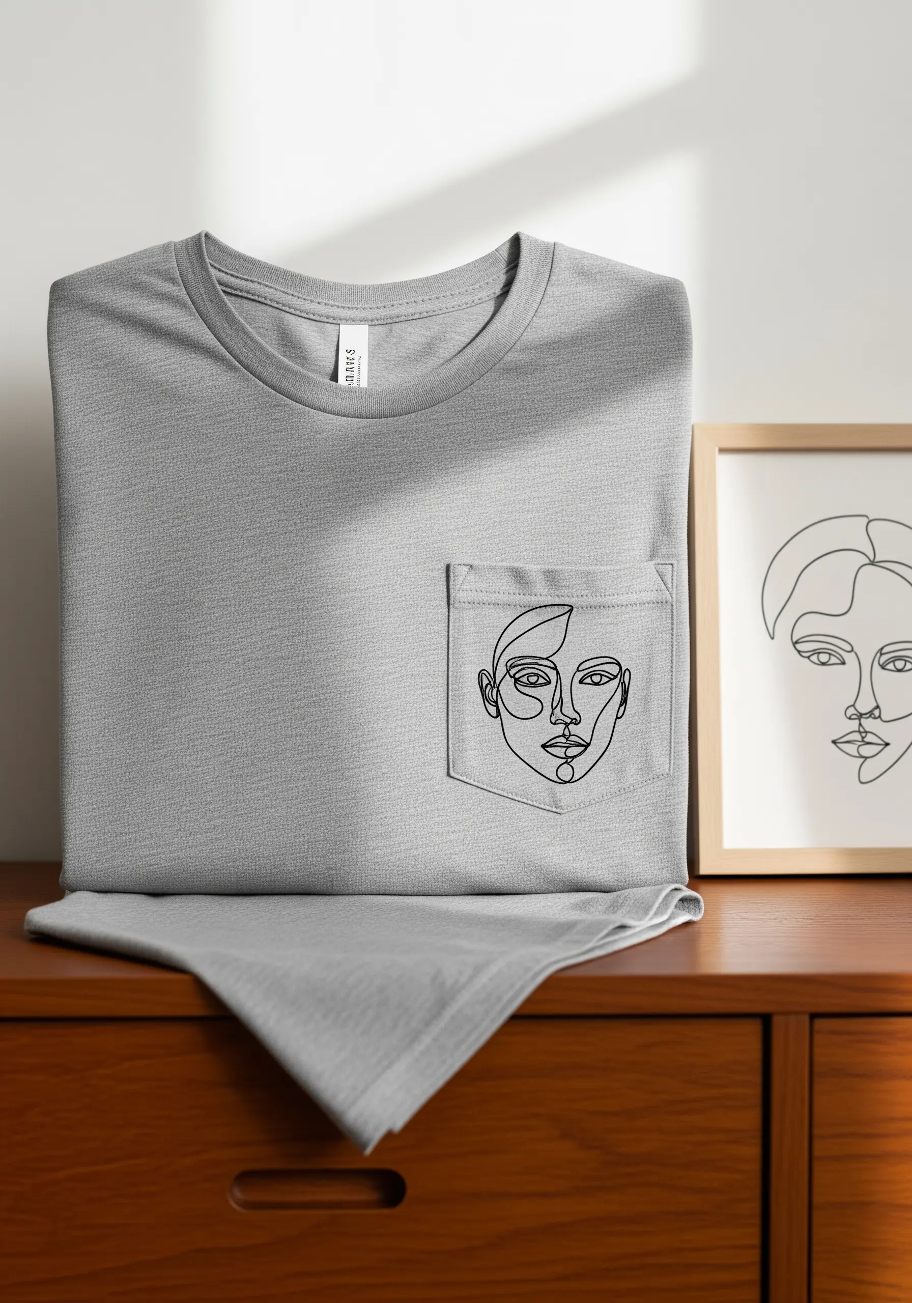 A gray t-shirt with a minimalist black-thread abstract face embroidered on the pocket.