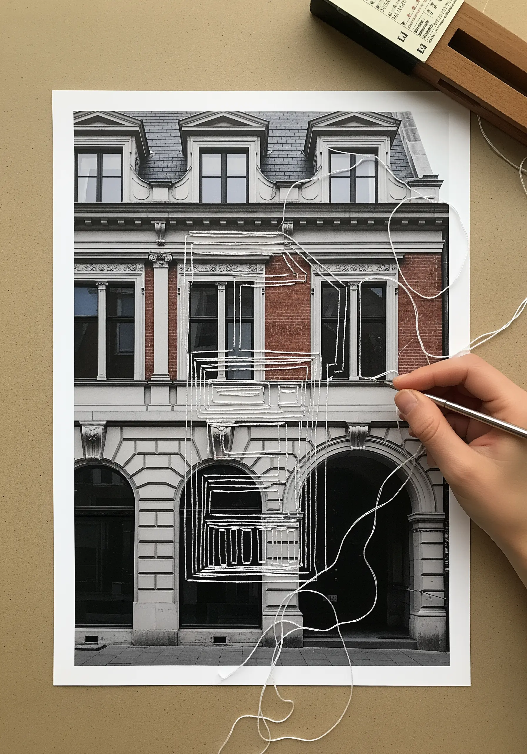 A hand stitching white thread outlines onto a black and white photograph of a building.