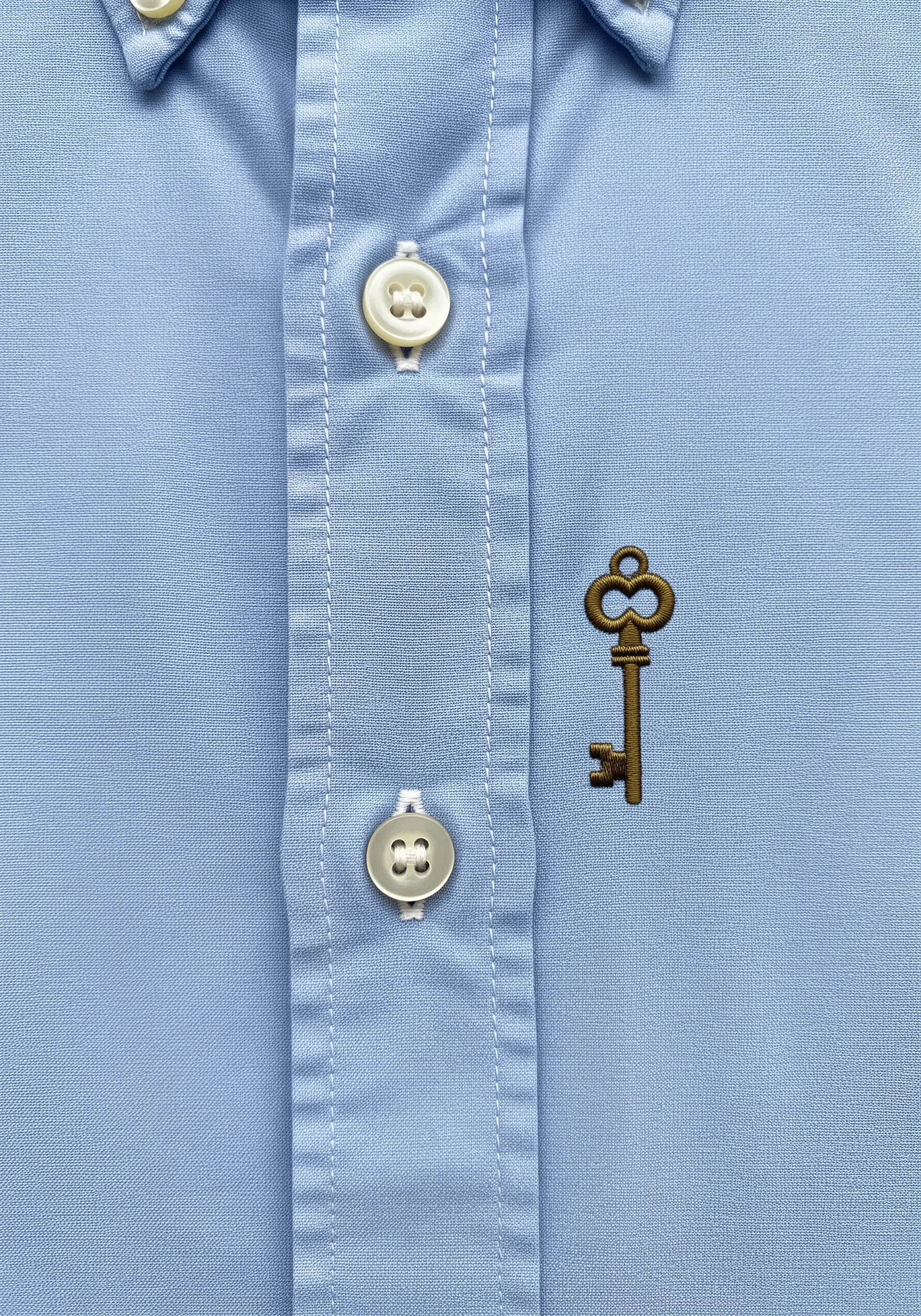 A small, detailed vintage key embroidered in bronze thread on the placket of a blue shirt.