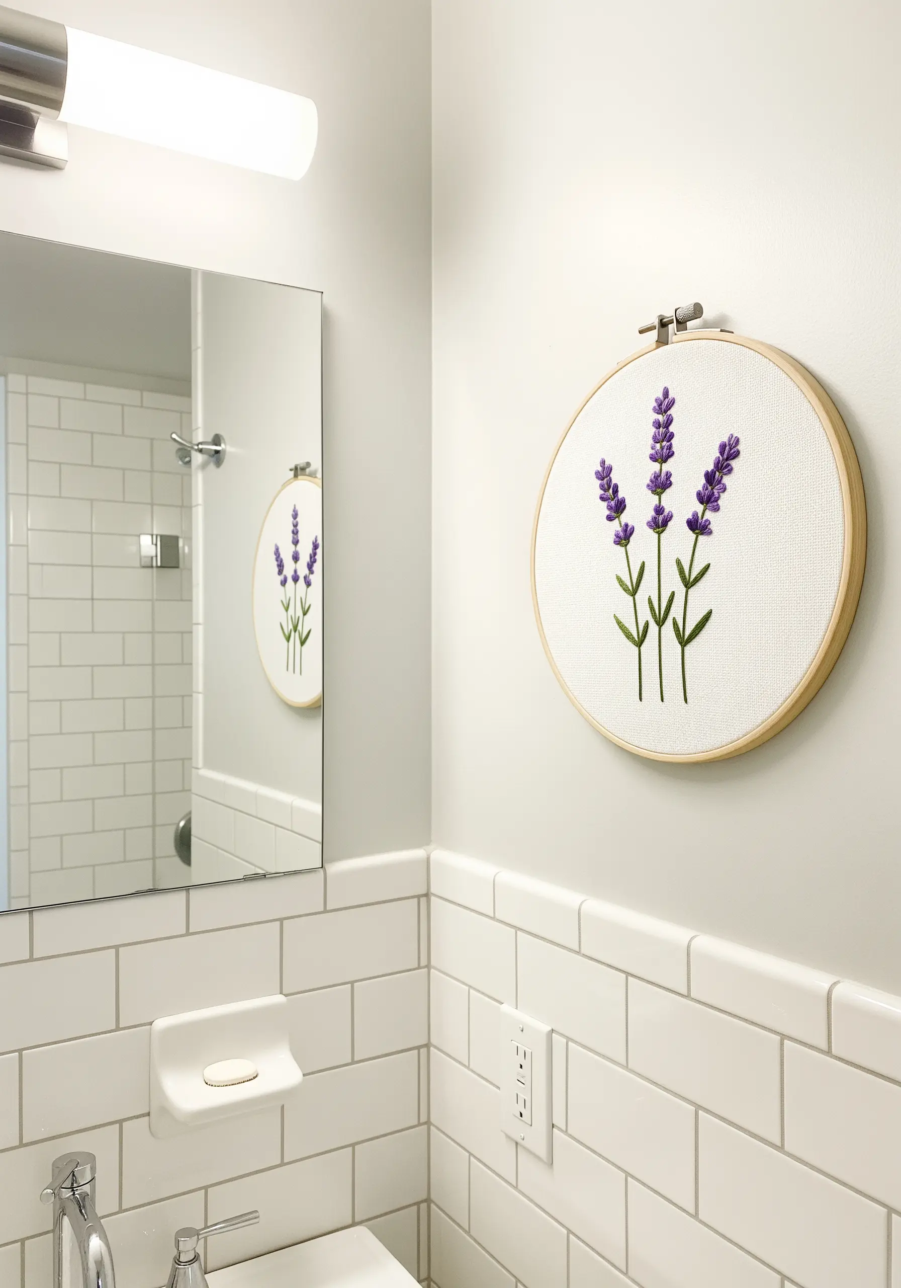 A simple embroidery of three lavender sprigs in a wooden hoop hanging in a white bathroom.