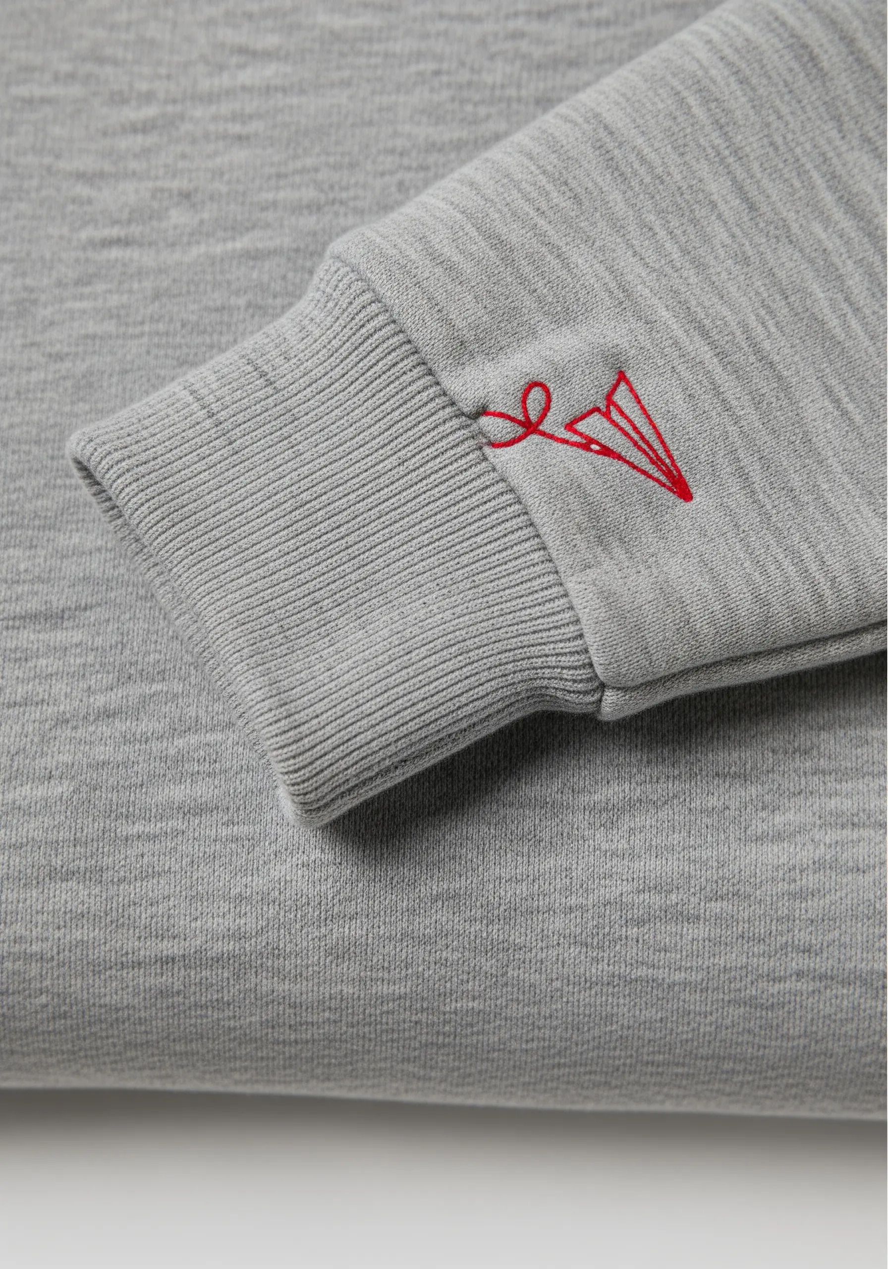 Tiny red paper airplane line art embroidered on a grey sweater cuff.