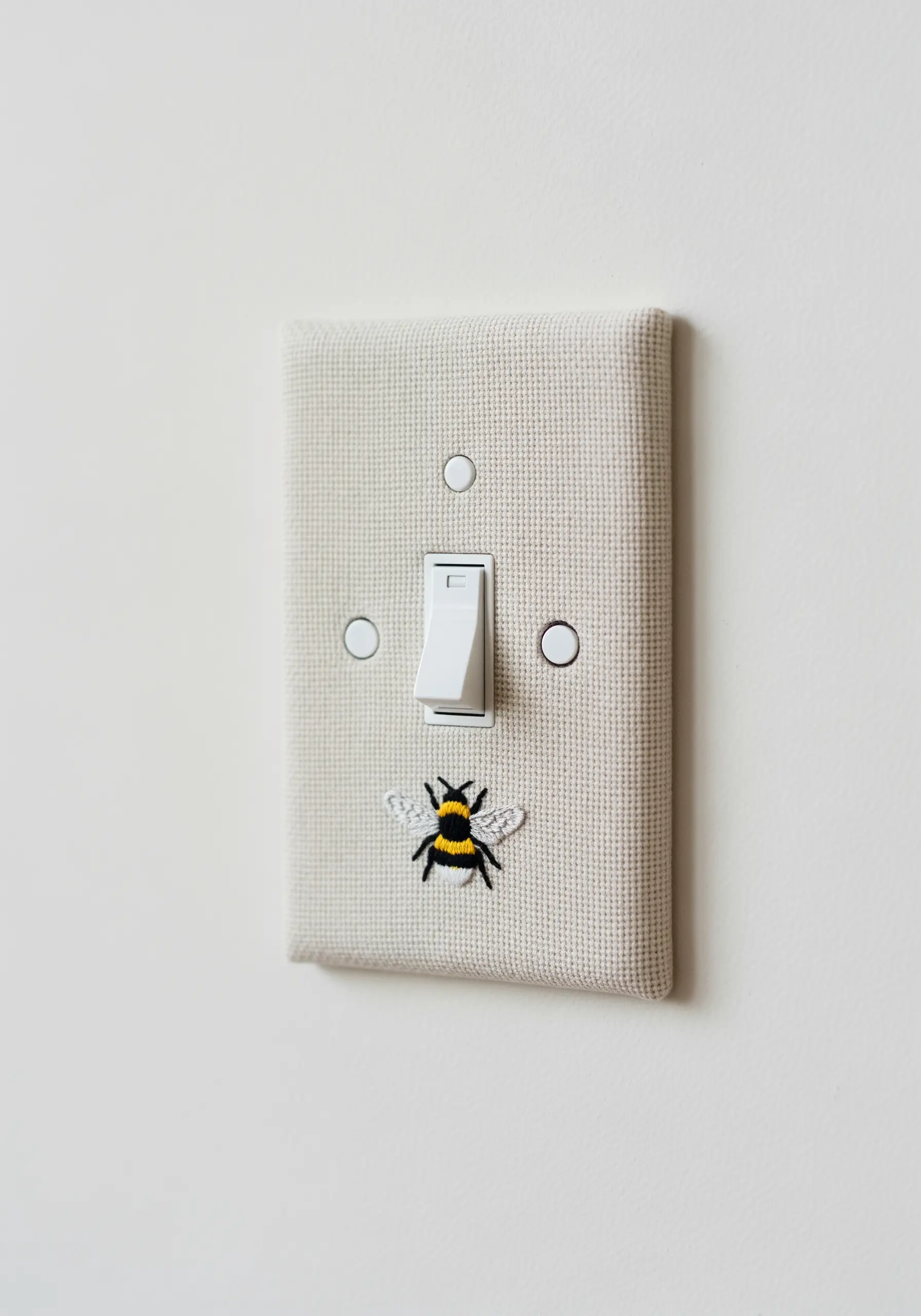 A fabric-covered light switch plate with a tiny, detailed embroidered bee at the bottom