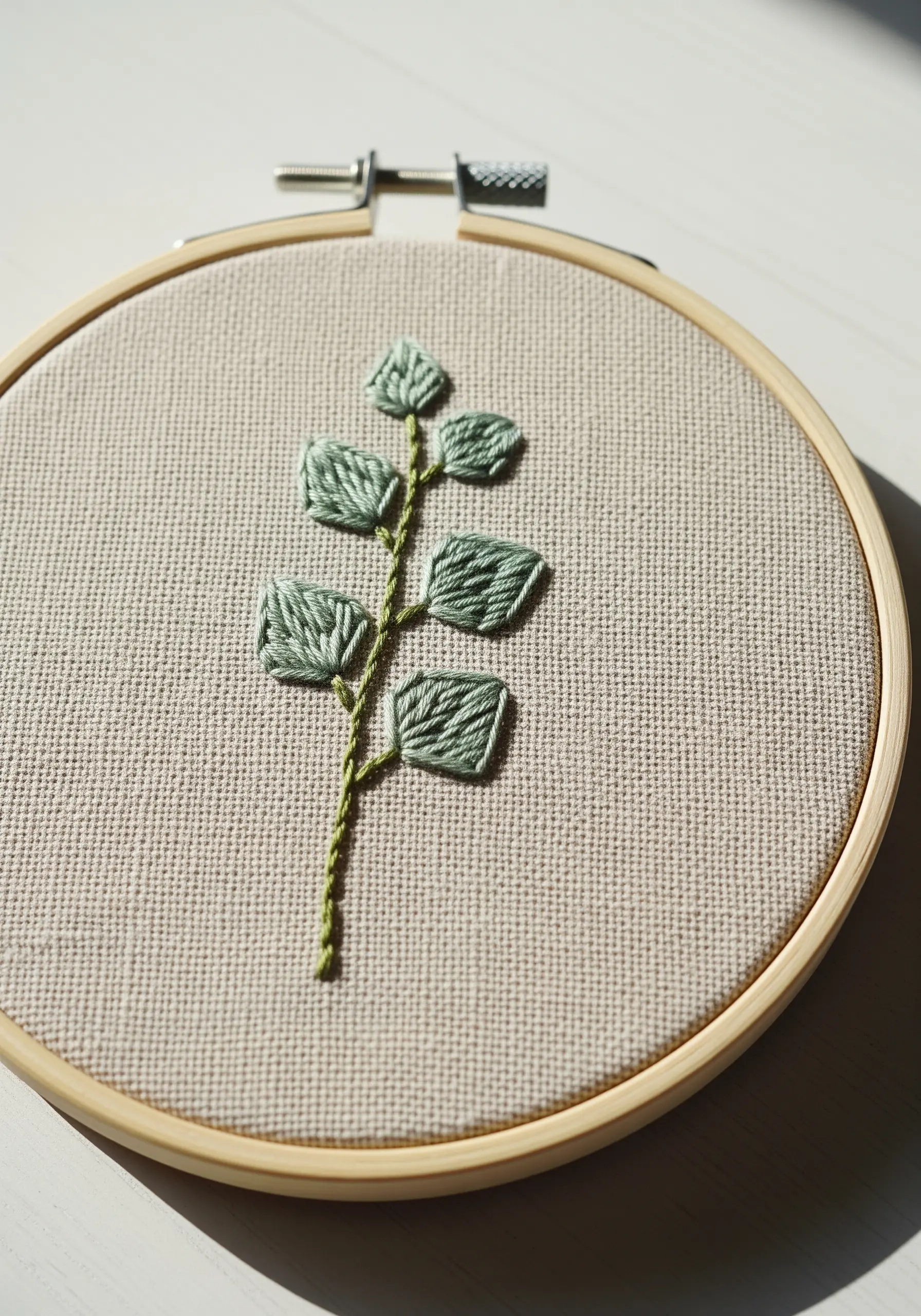 A simple embroidered branch with sage green leaves on neutral linen fabric.