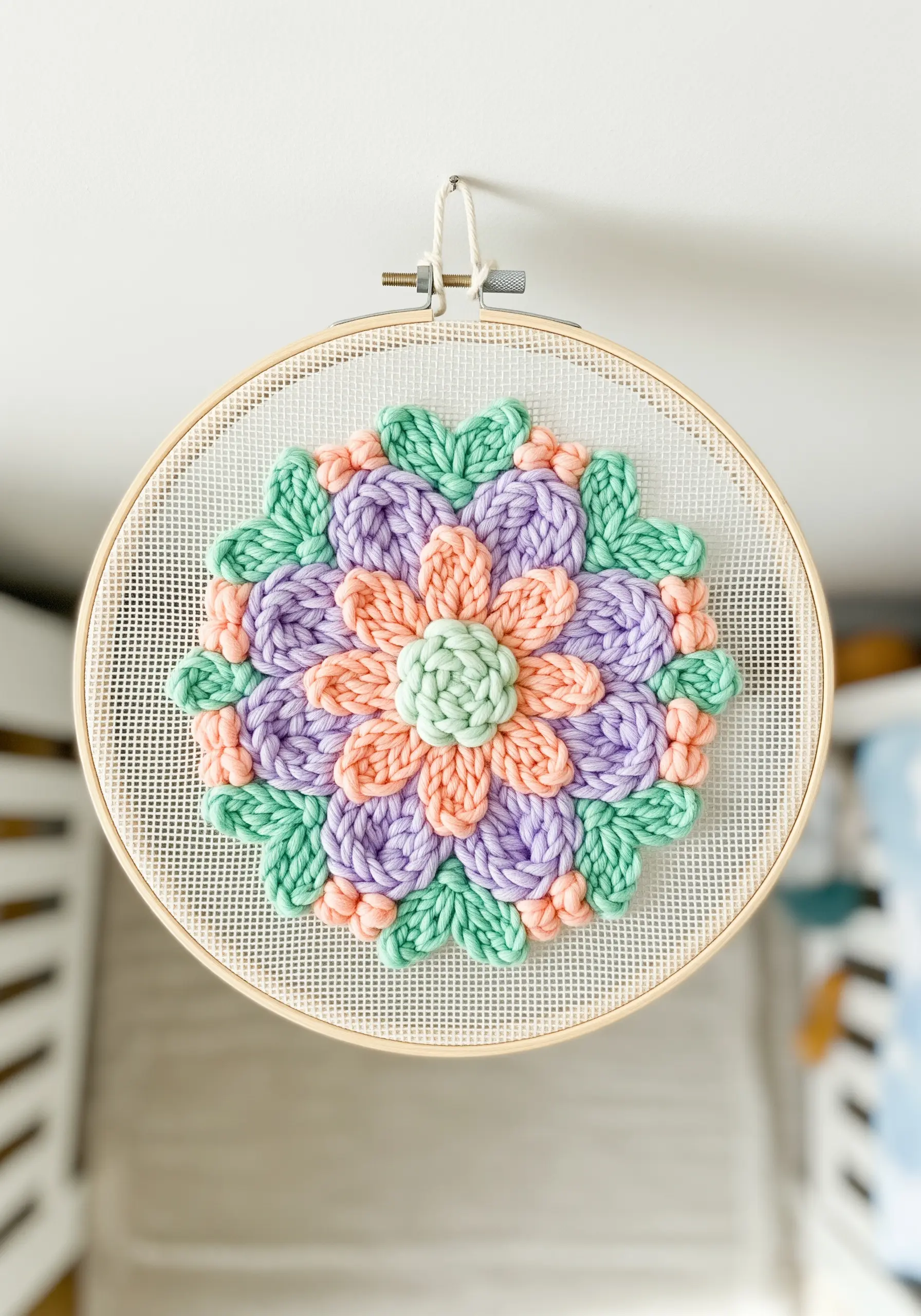 A dimensional punch needle flower with raised petals in pastel colors, hung in a nursery.