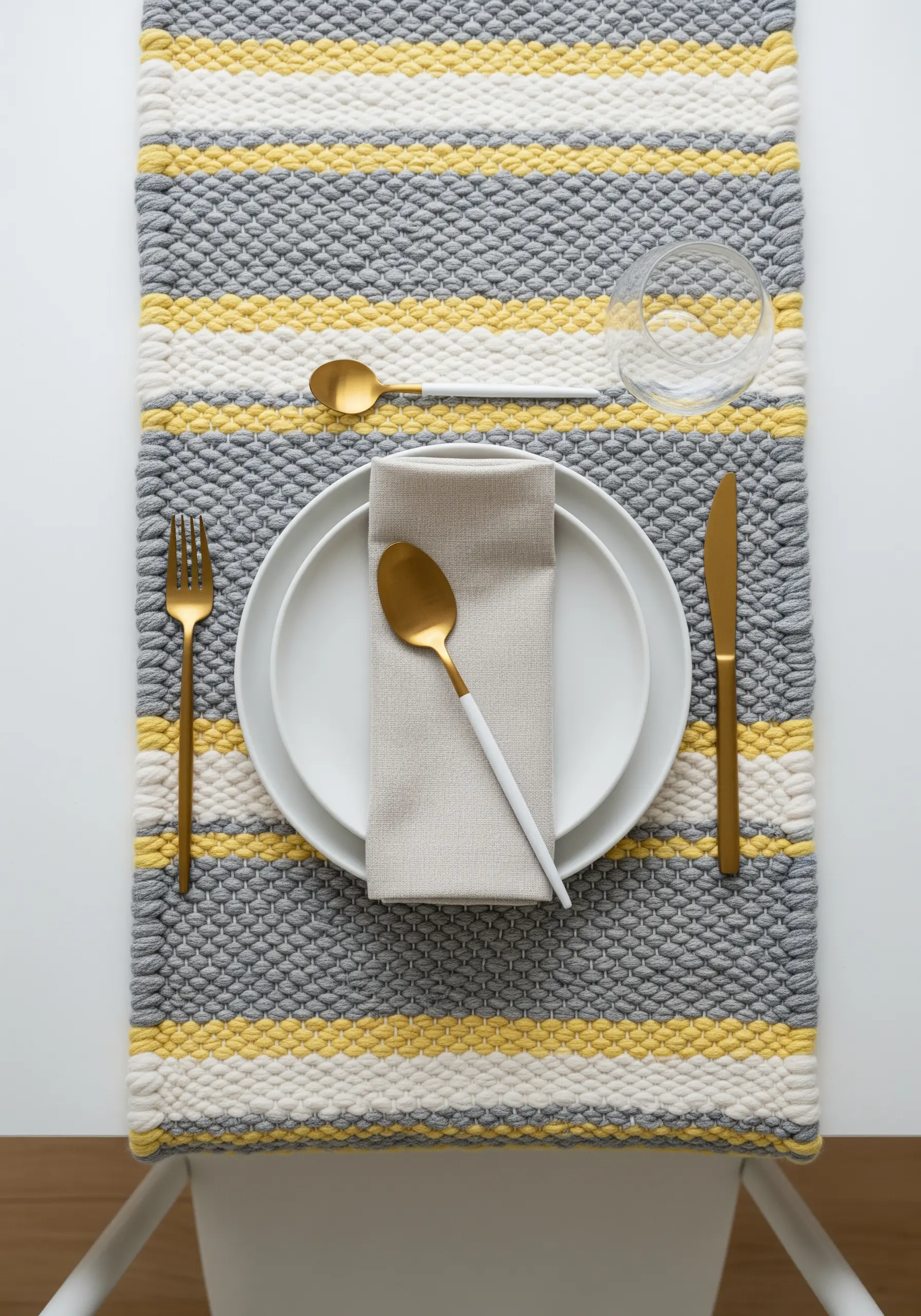 A woven grey, yellow, and white table runner set with a white plate and gold flatware.
