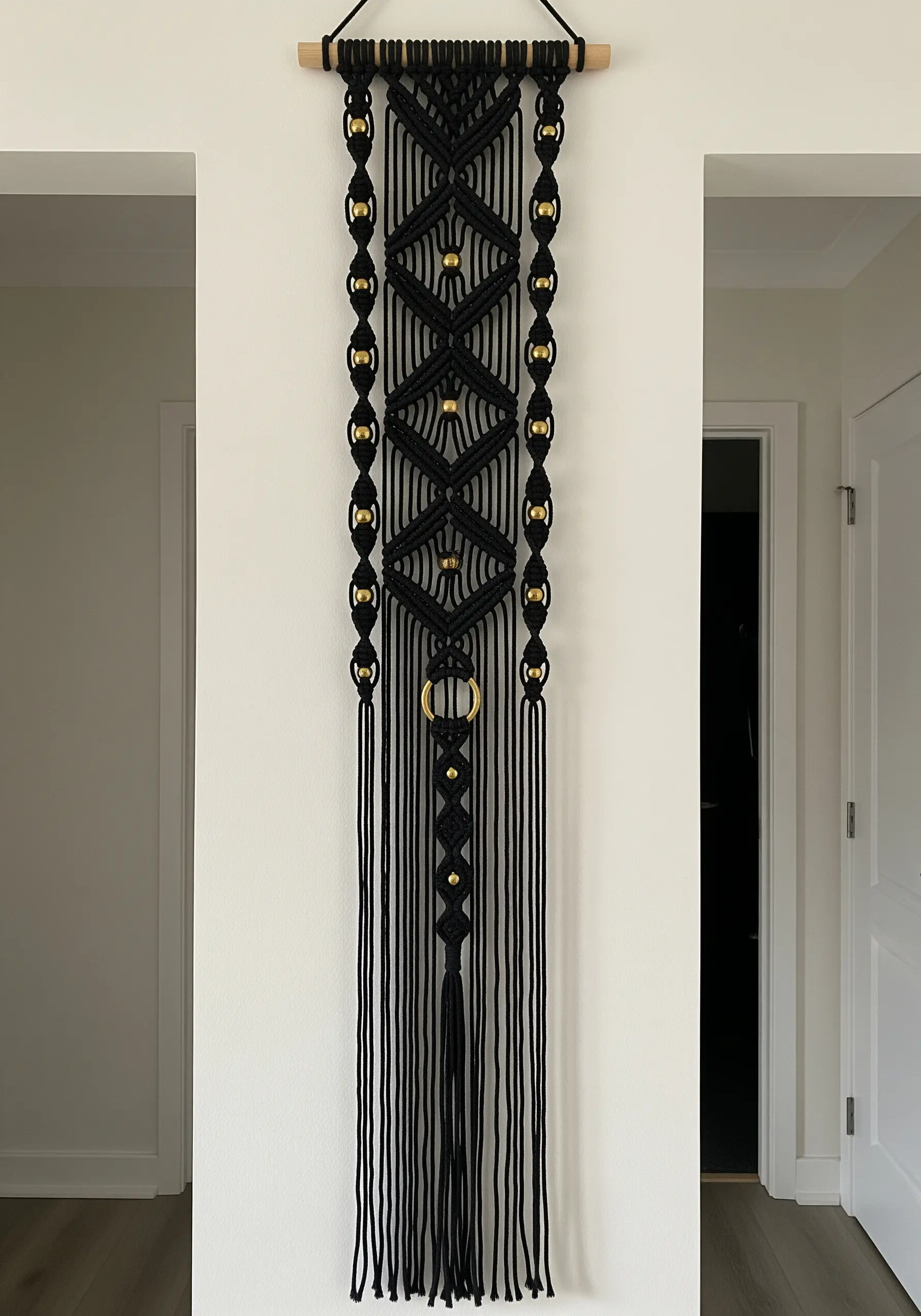 A long, thin black macrame wall hanging adorned with gold beads and a central gold ring