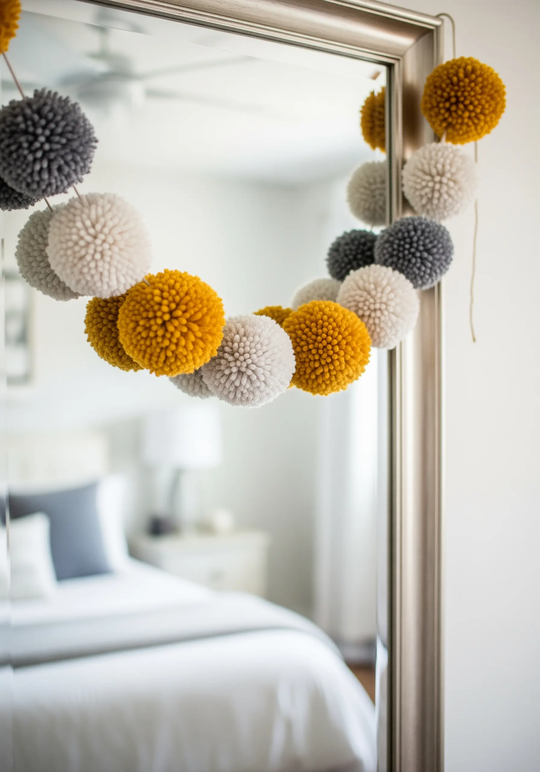 A fluffy pom-pom garland in mustard yellow, grey, and off-white hanging on a mirror.