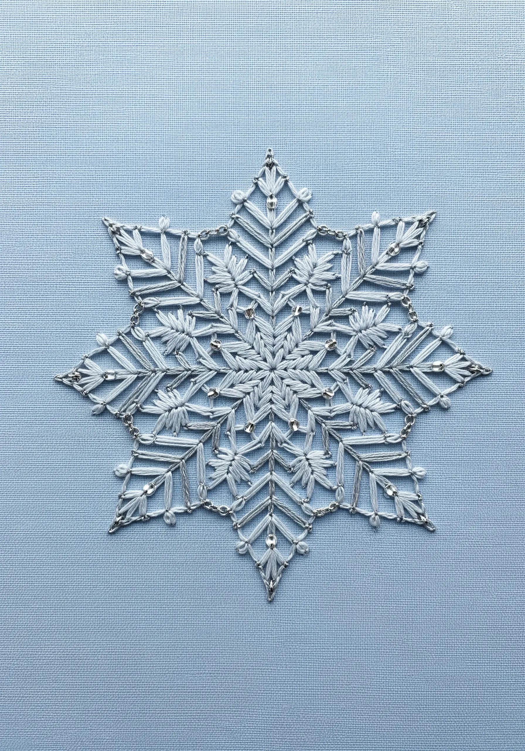 An intricate white and silver snowflake mandala with tiny beads on light blue fabric.