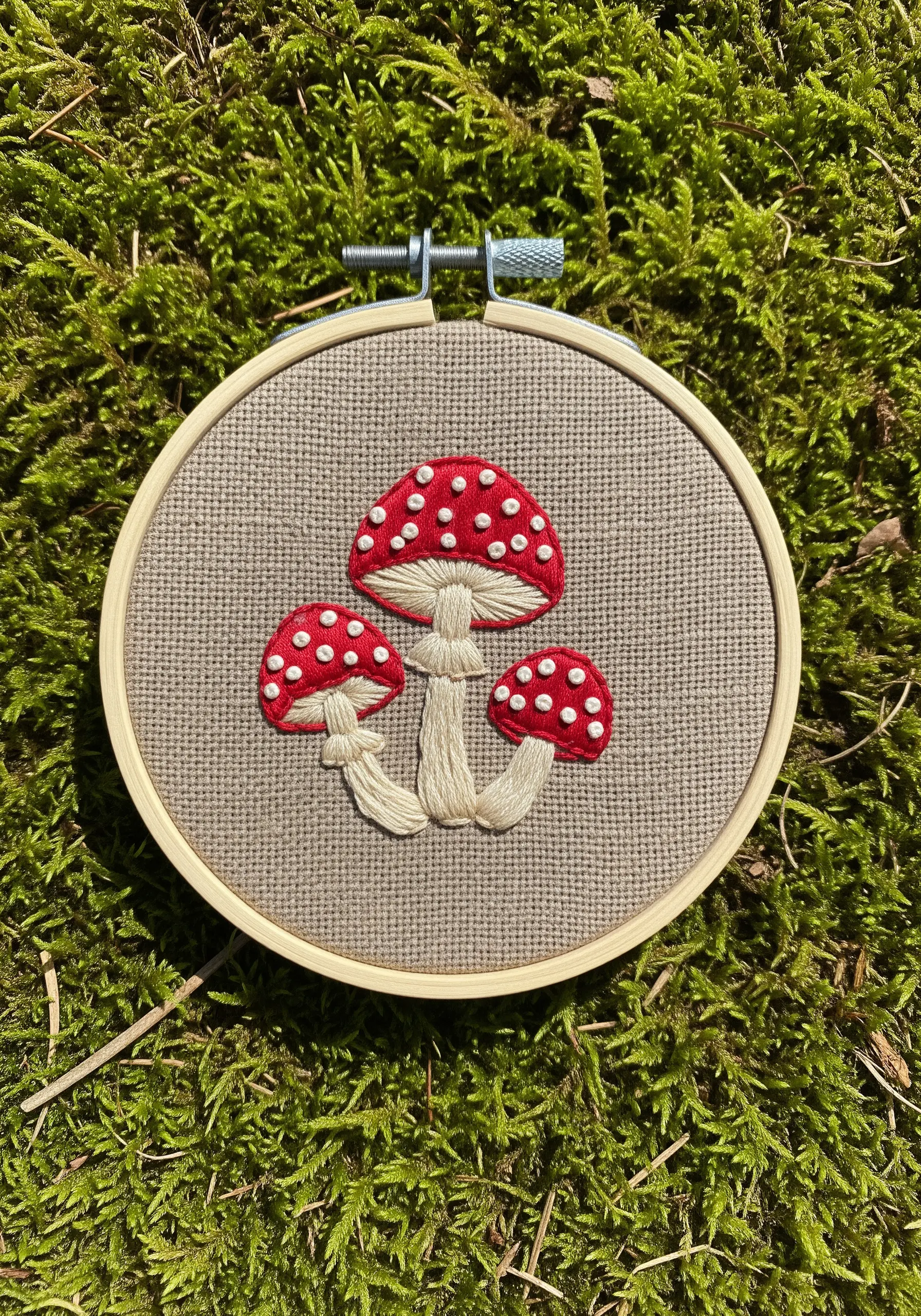 Three embroidered red toadstool mushrooms with white French knot spots on linen fabric.