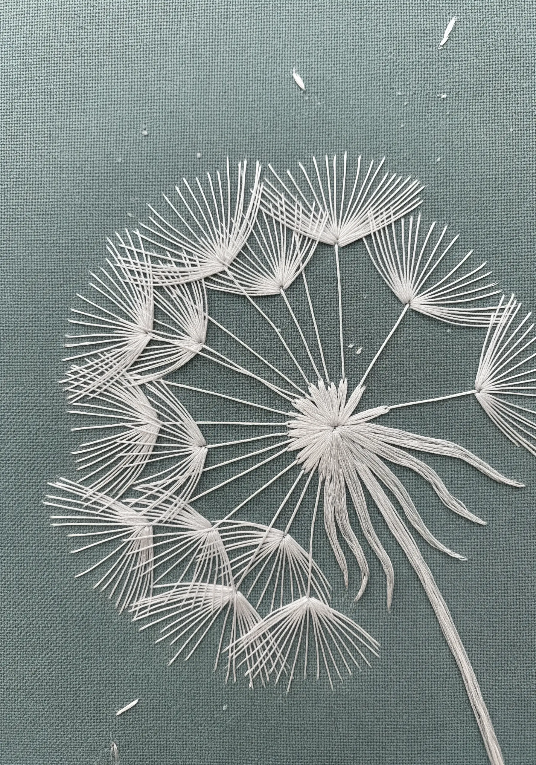 A delicate white dandelion seed head on blue-grey fabric using straight stitches.