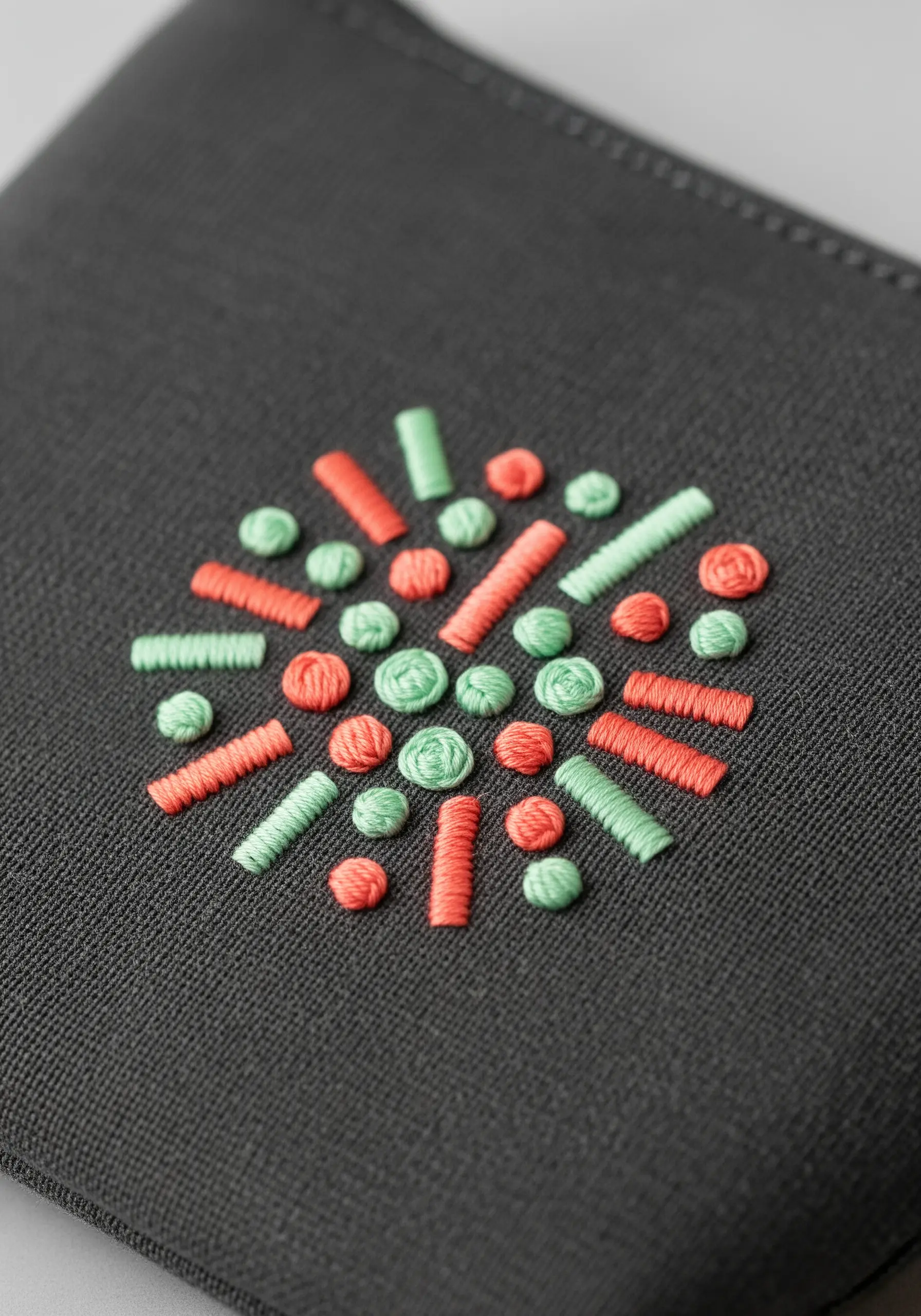 An abstract circular pattern of small stitches in coral and mint on a dark pouch.