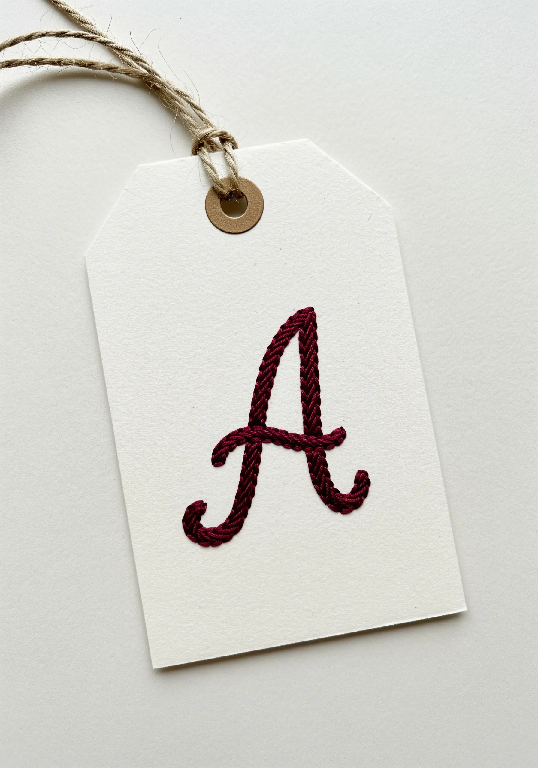 A bold, dark red embroidered letter 'A' on a cream paper gift tag.