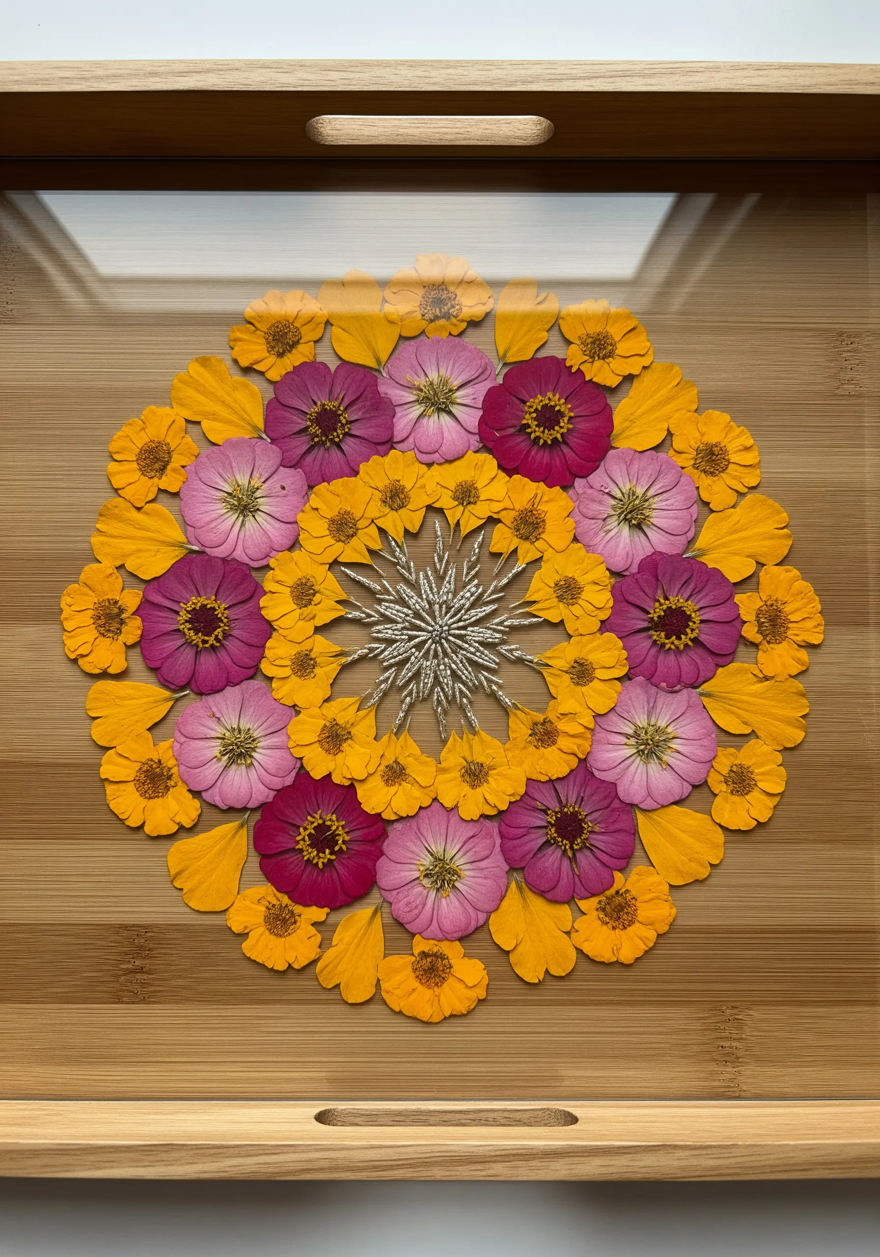 A mandala of pressed flower petals on a wooden tray with an embroidered silver centerpiece.