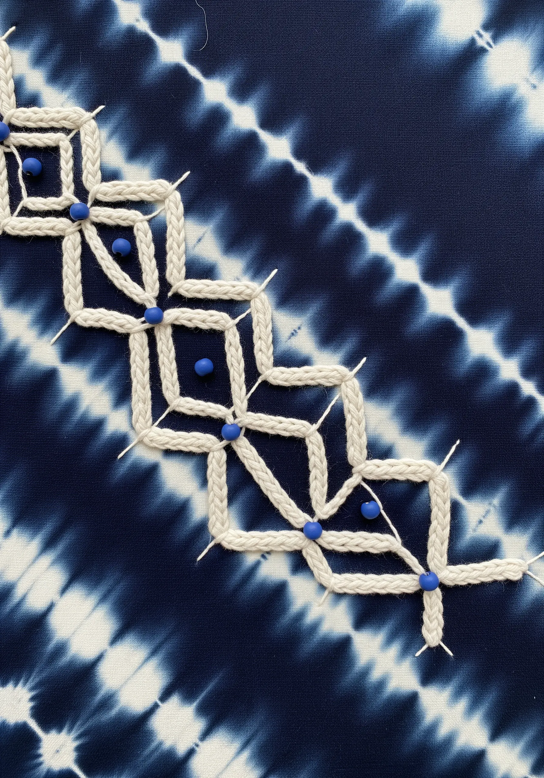A geometric macrame-style pattern on indigo tie-dye fabric, with blue beads at the intersections.