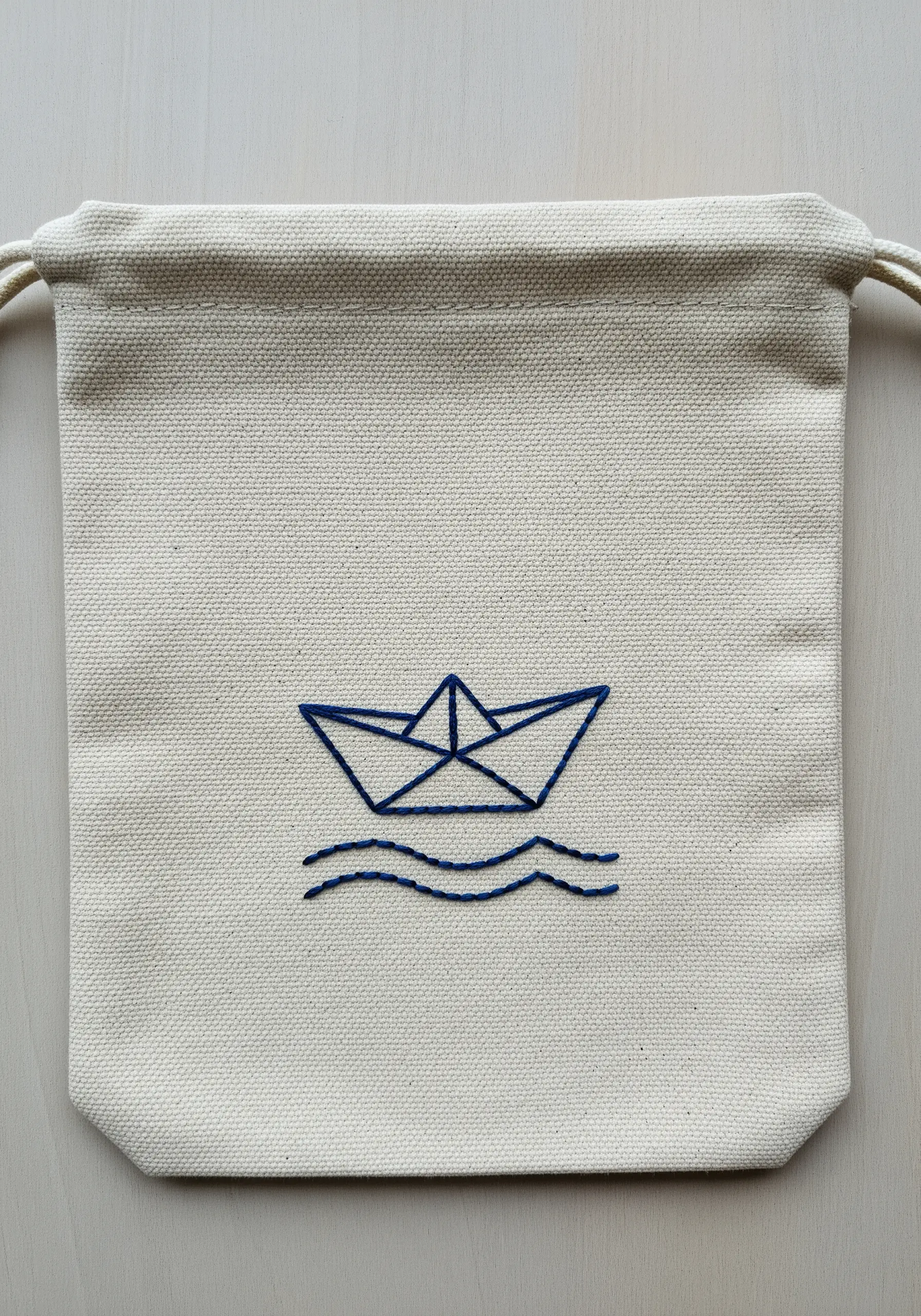 A simple outline of a paper boat and waves embroidered in blue on a canvas pouch.