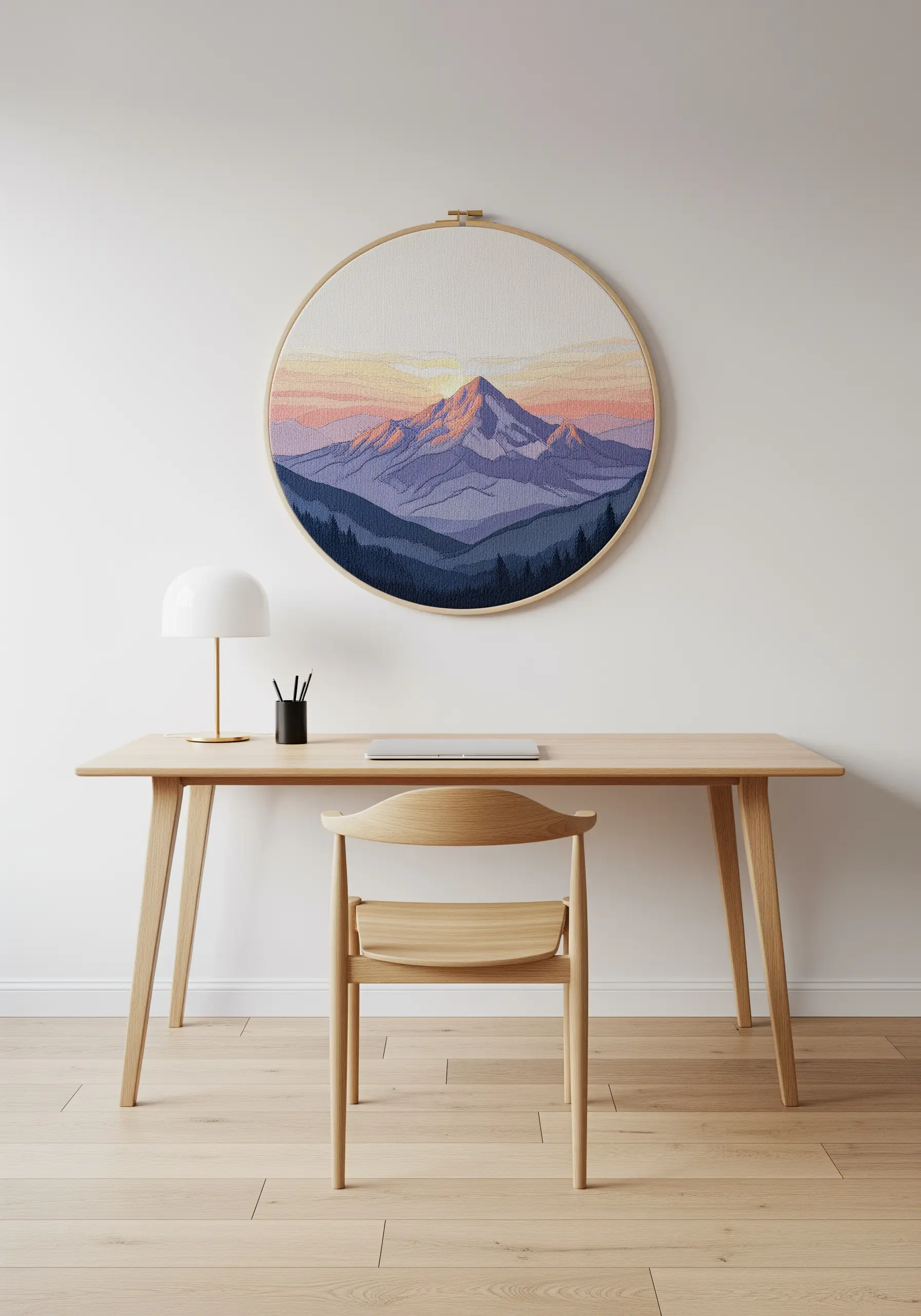 Large embroidery hoop with a thread-painted mountain landscape at sunset, hanging above a desk.