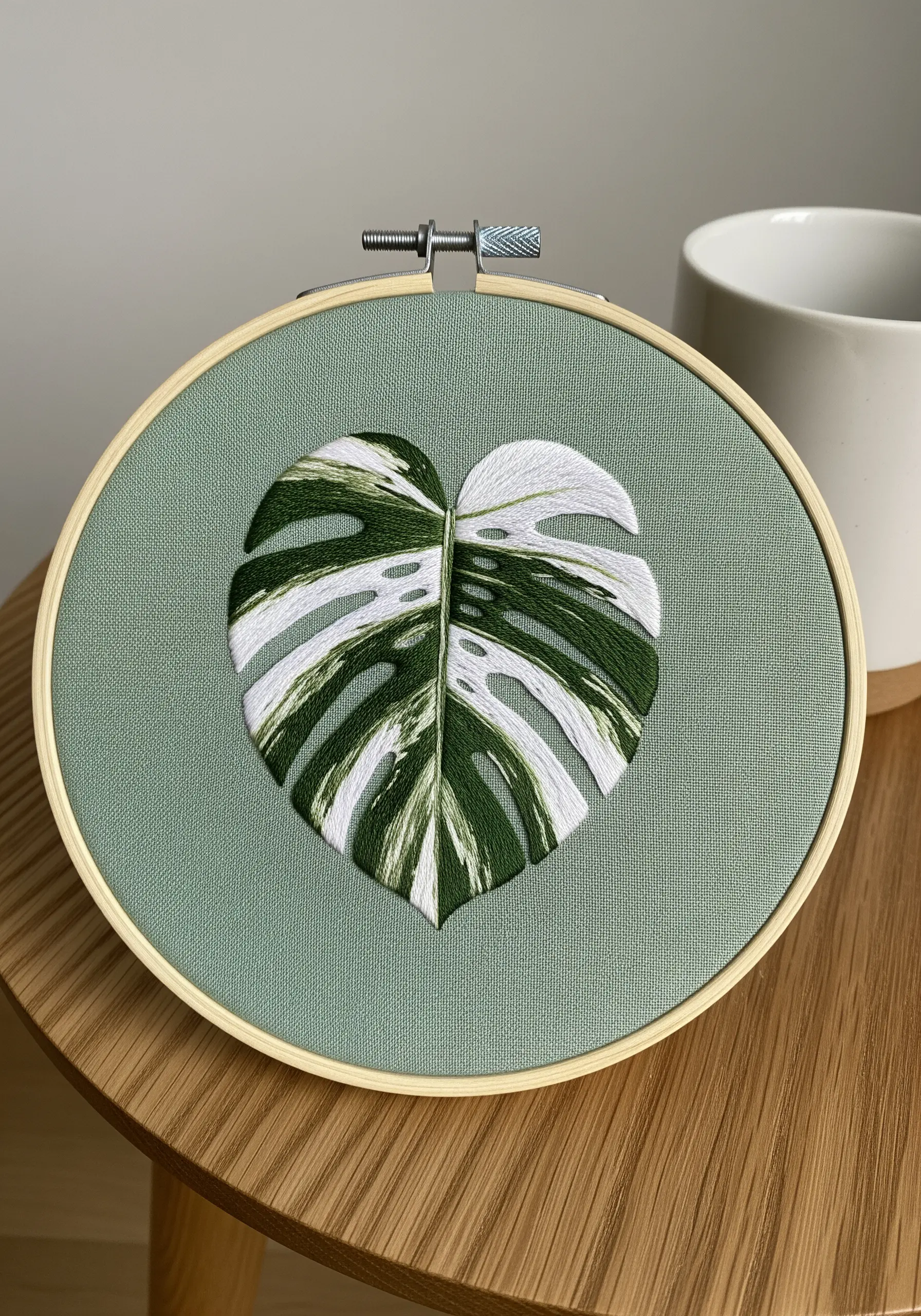 A variegated Monstera leaf with white and green sections stitched on sage green fabric.