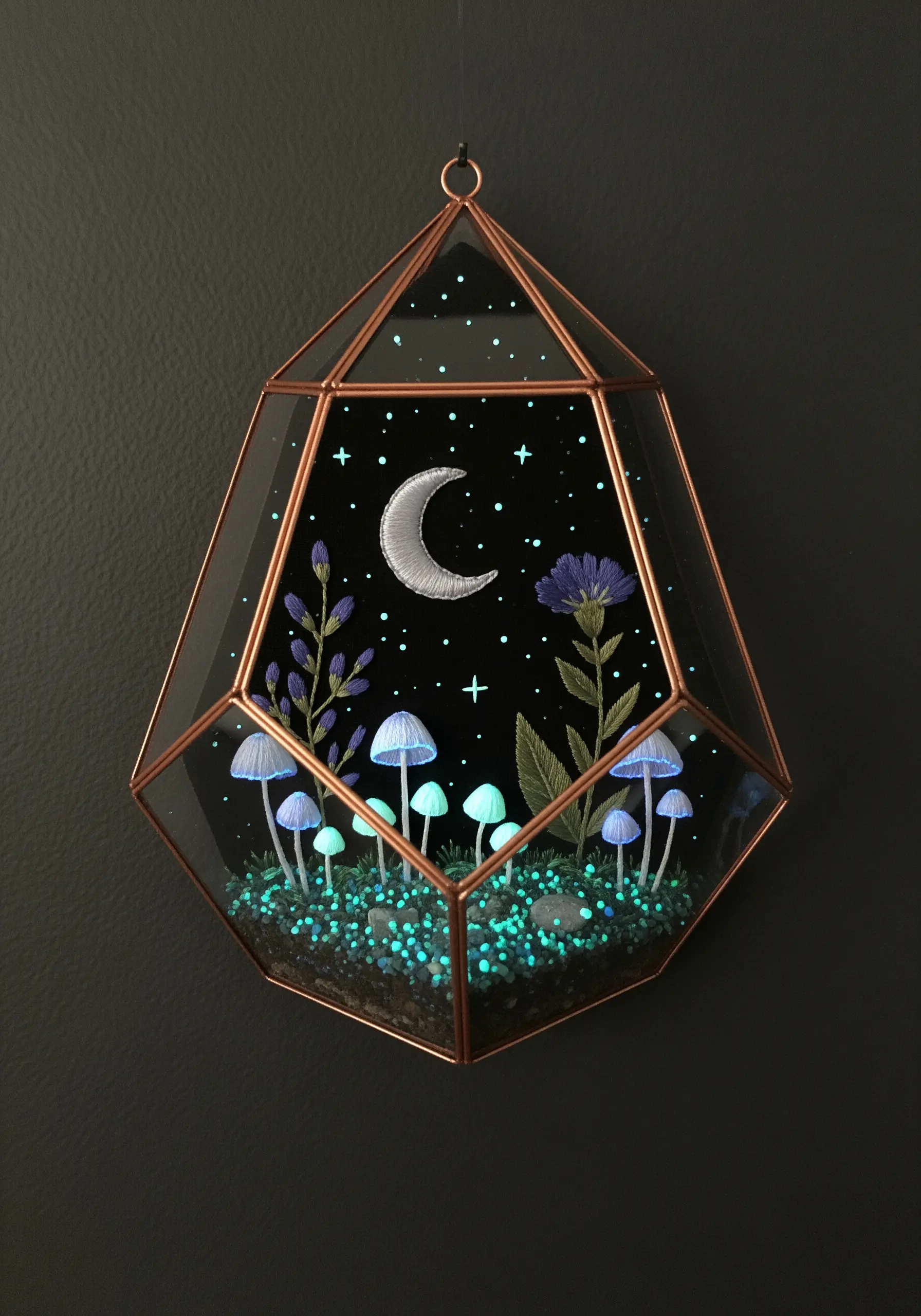 An embroidered night scene with mushrooms and a moon inside a geometric glass terrarium.
