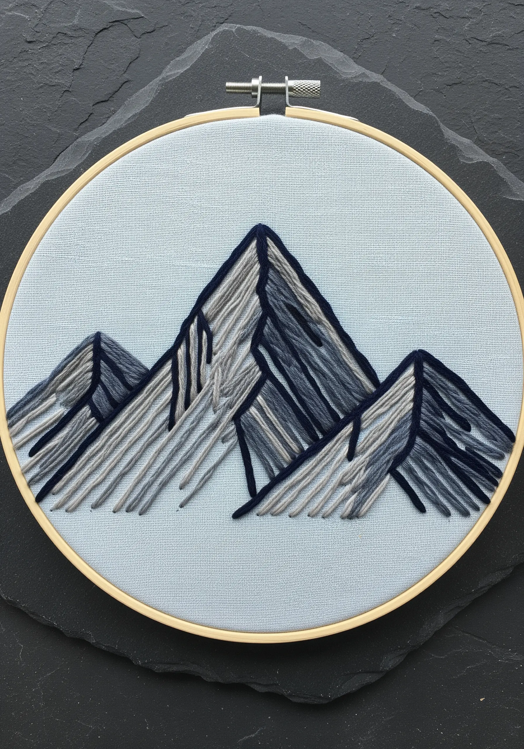 A minimalist embroidery of mountain peaks using blocks of grey and blue straight stitches.