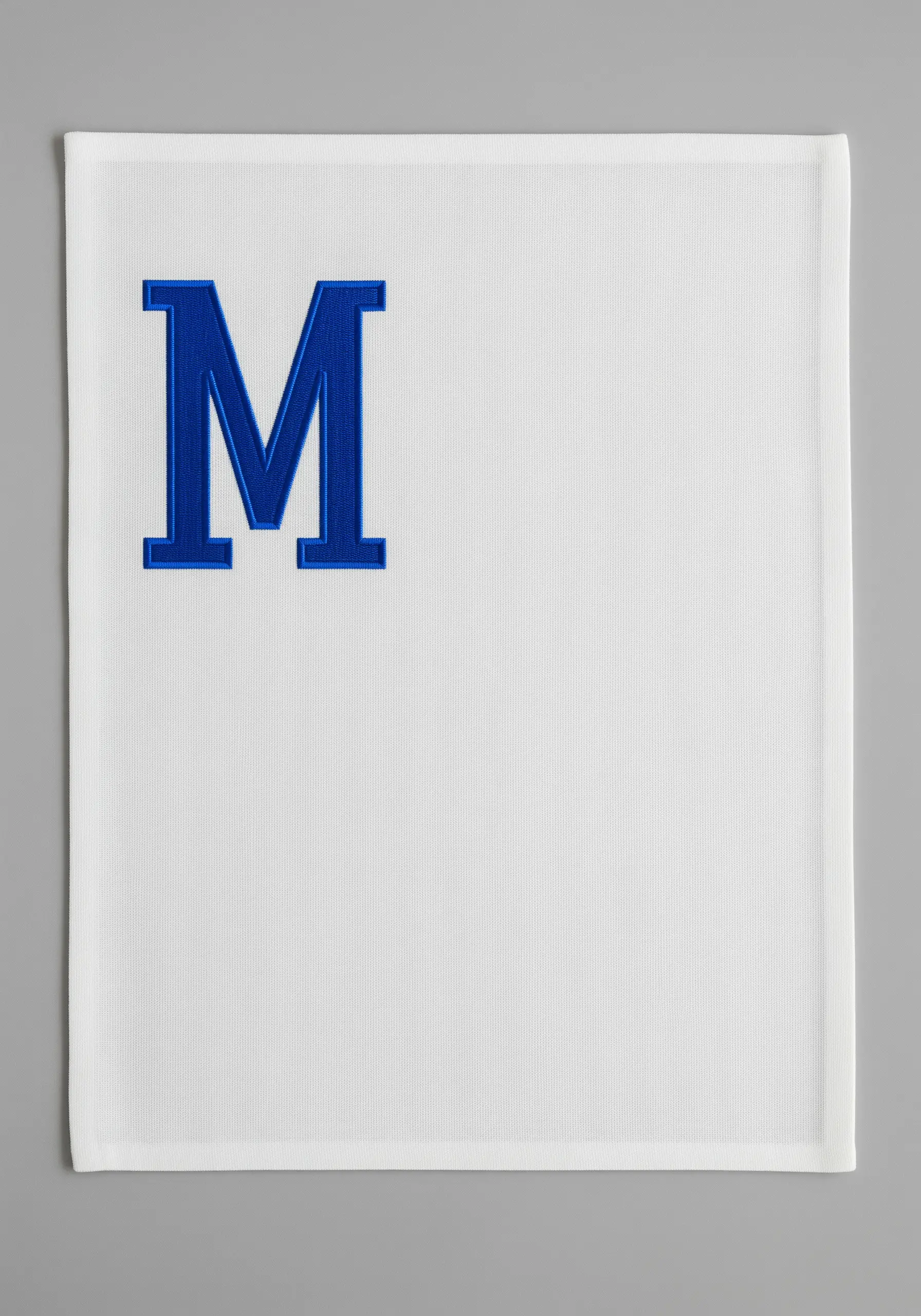 A single, bold, royal blue satin-stitched block letter 'M' on a white piqué napkin.