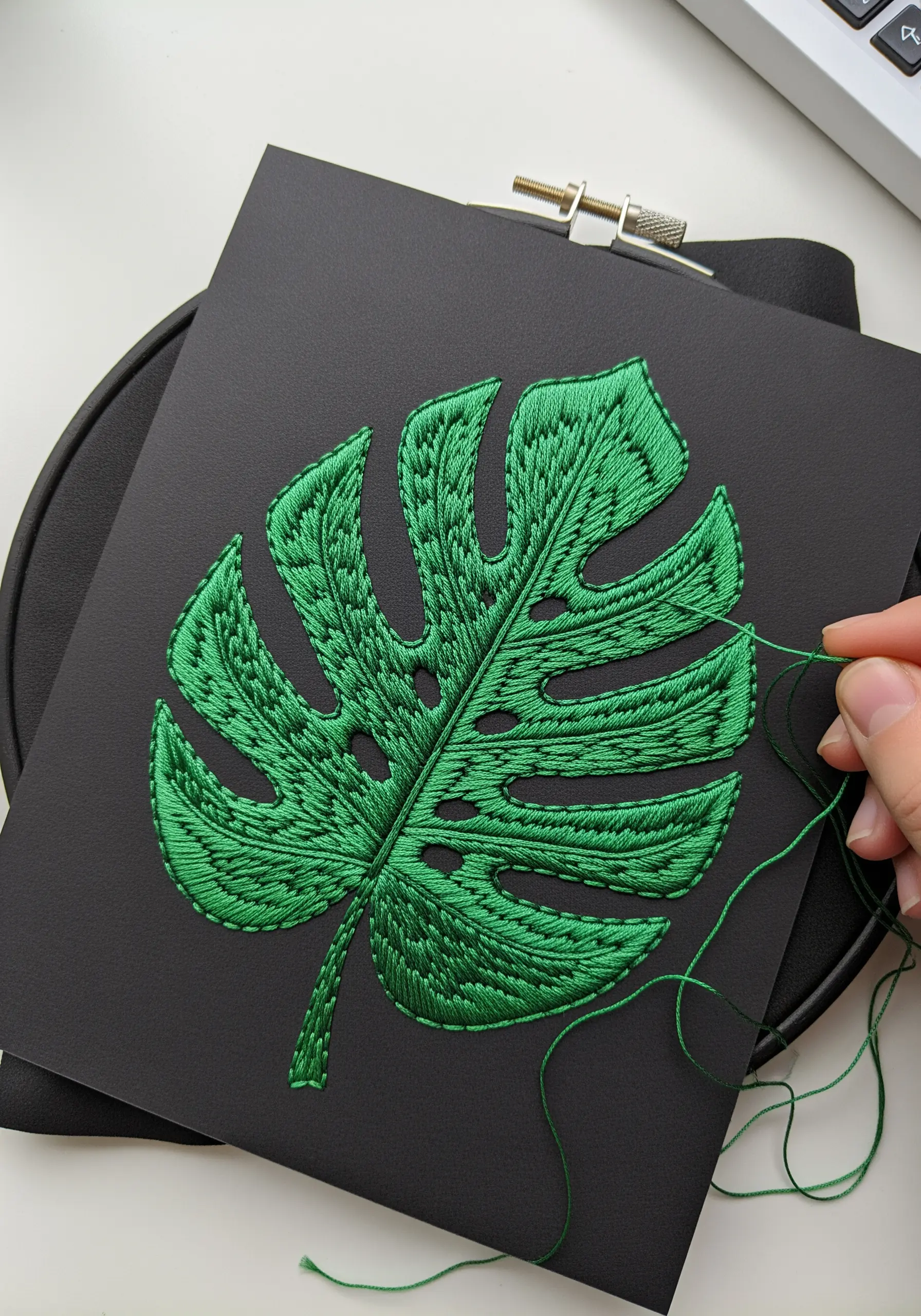 A vibrant green satin-stitched monstera leaf embroidered on black cardstock.