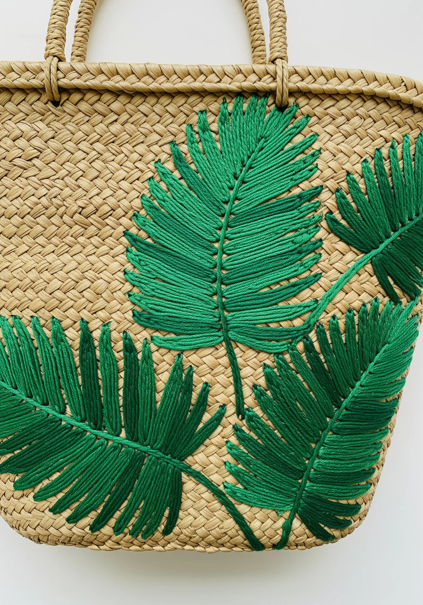 Large, vibrant green palm leaves embroidered with thick thread onto a woven straw bag.
