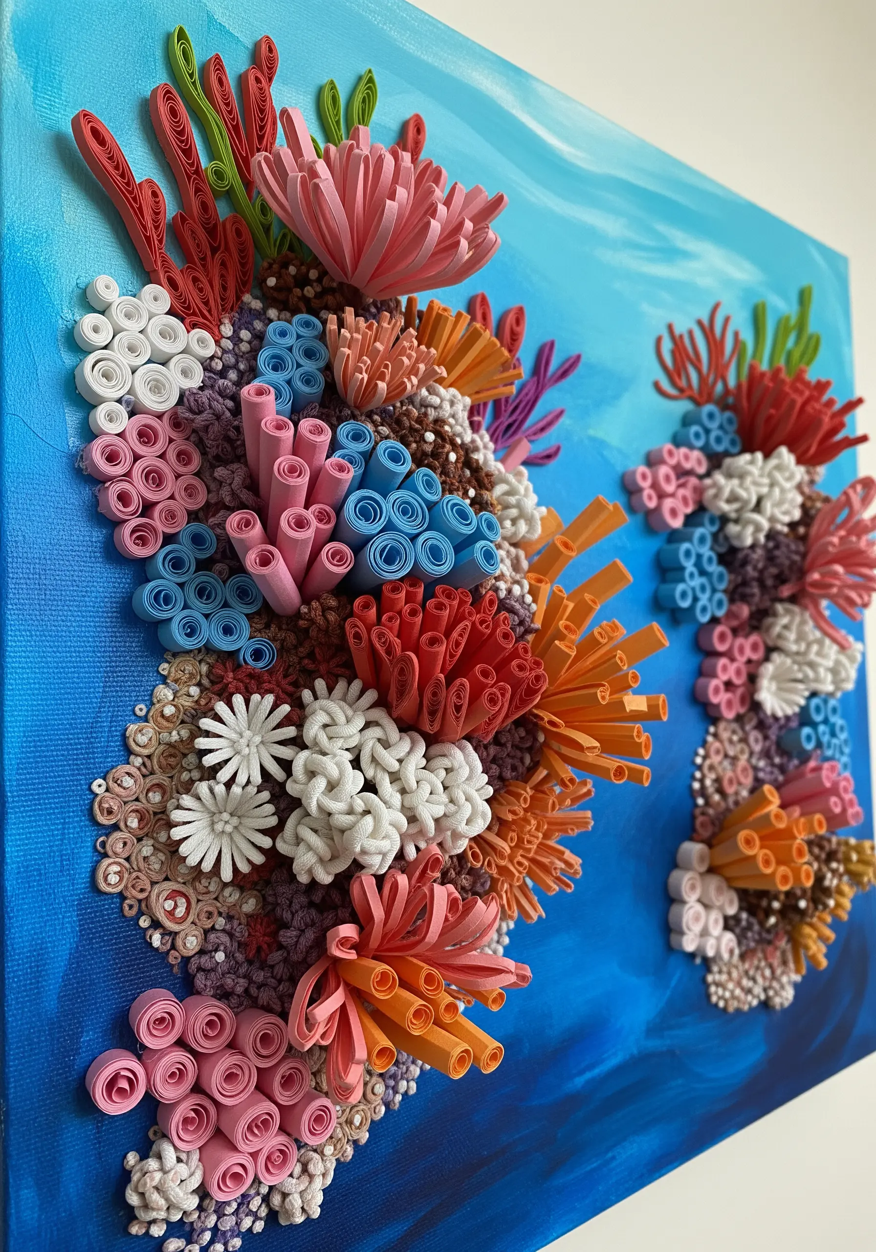 A vibrant 3D coral reef made from rolled and shaped paper strips on a blue painted canvas.