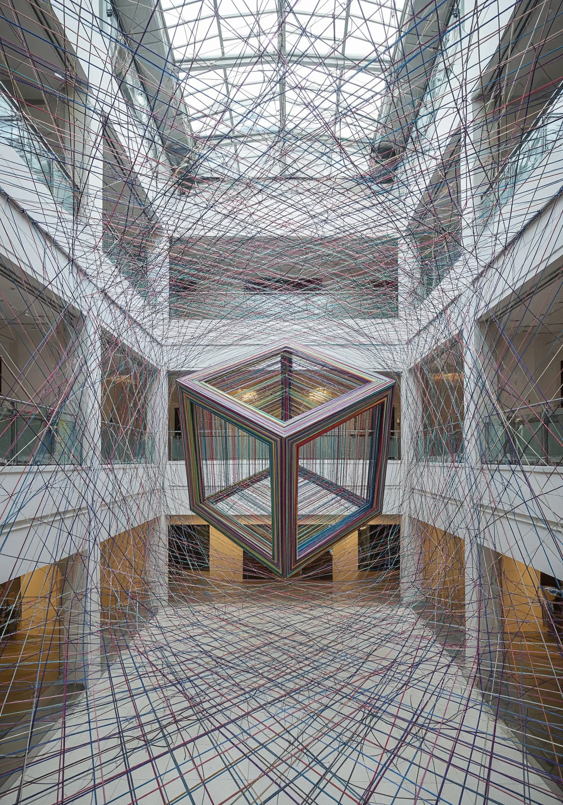 A multi-colored cube made of stretched threads appears to float in the center of a large atrium.
