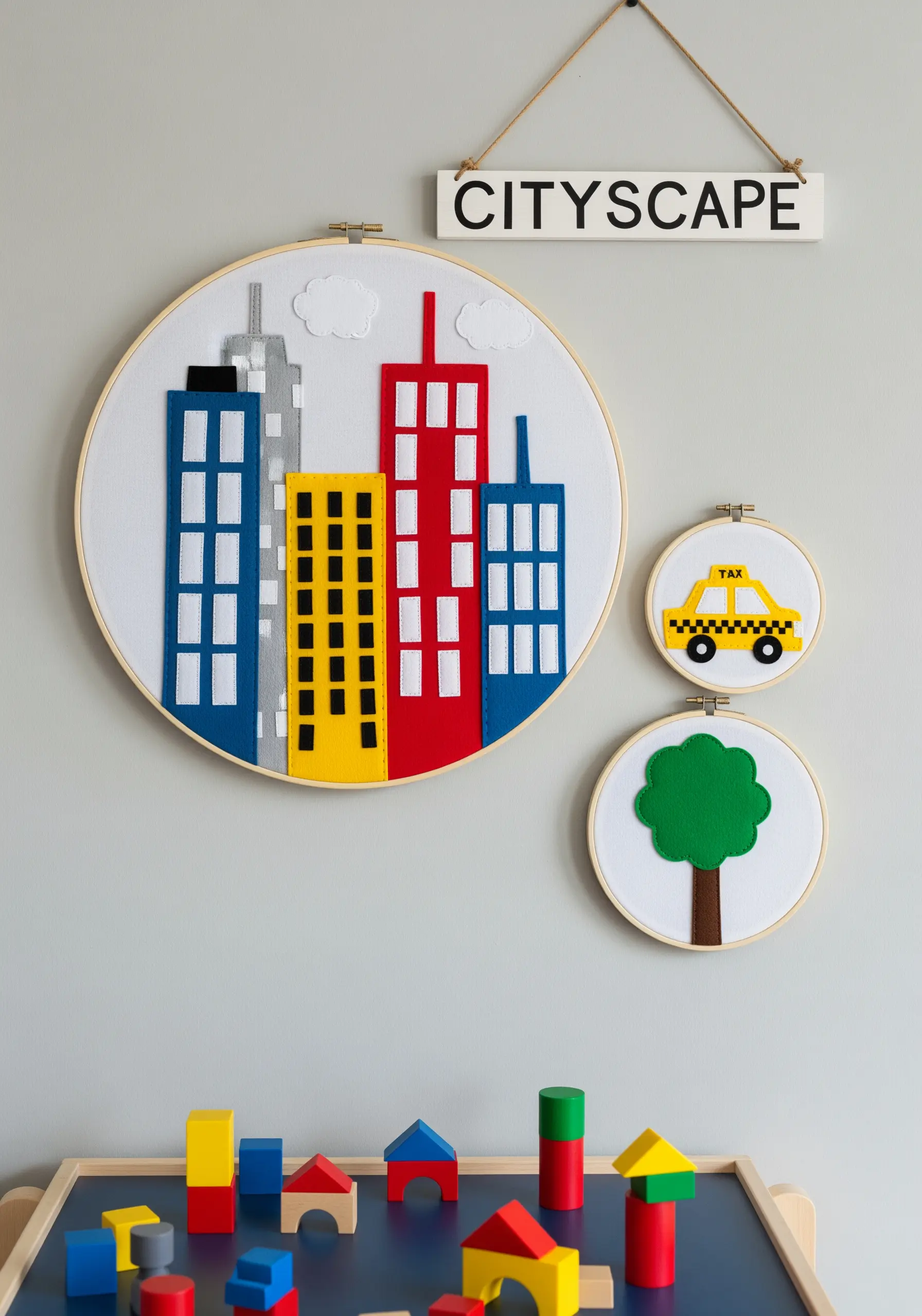 A large embroidery hoop with a colorful cityscape made from layered felt, next to smaller hoops with a car and tree.